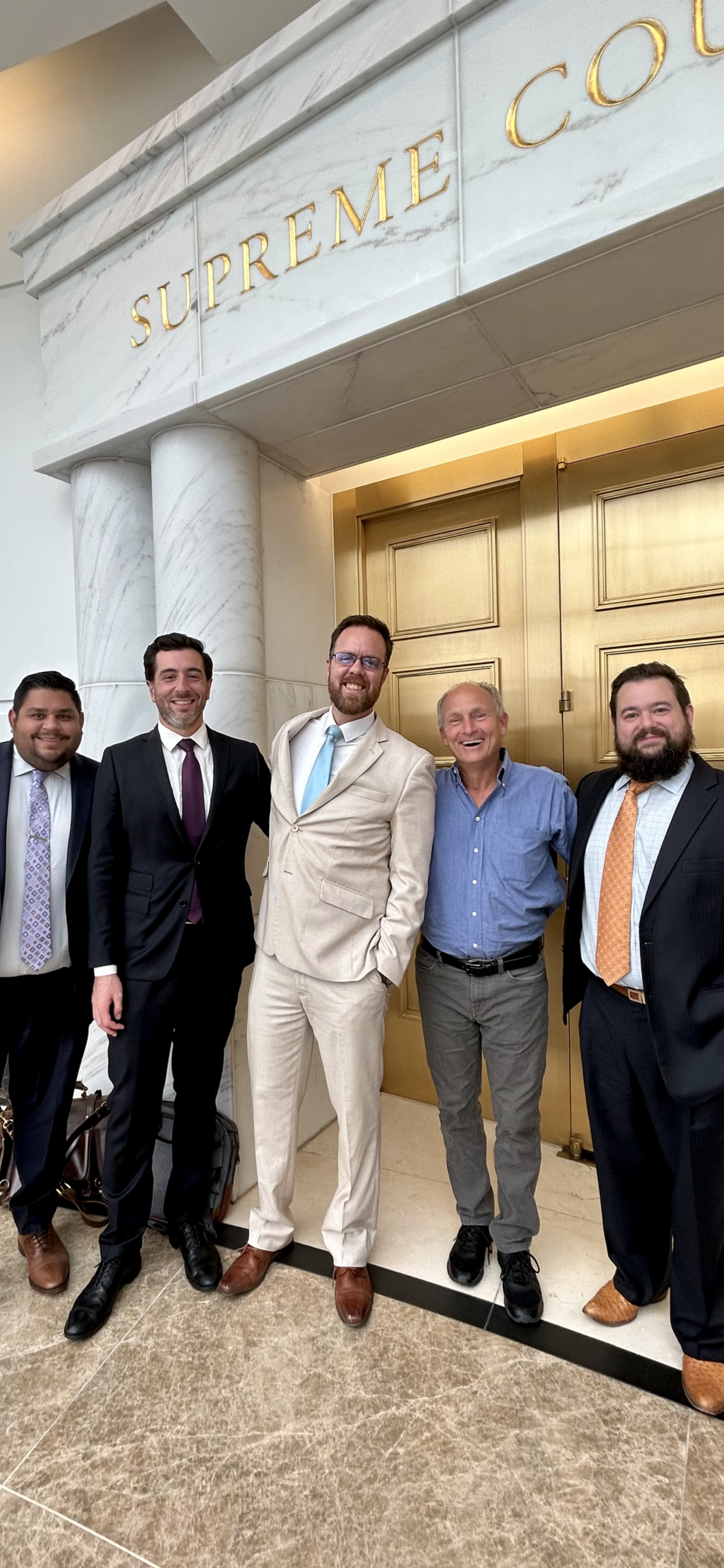 Five men dressed in suits standing together in front of a large gold door with a sign above that reads "Supreme Court" in gold letters. They are smiling and posing for the photo.