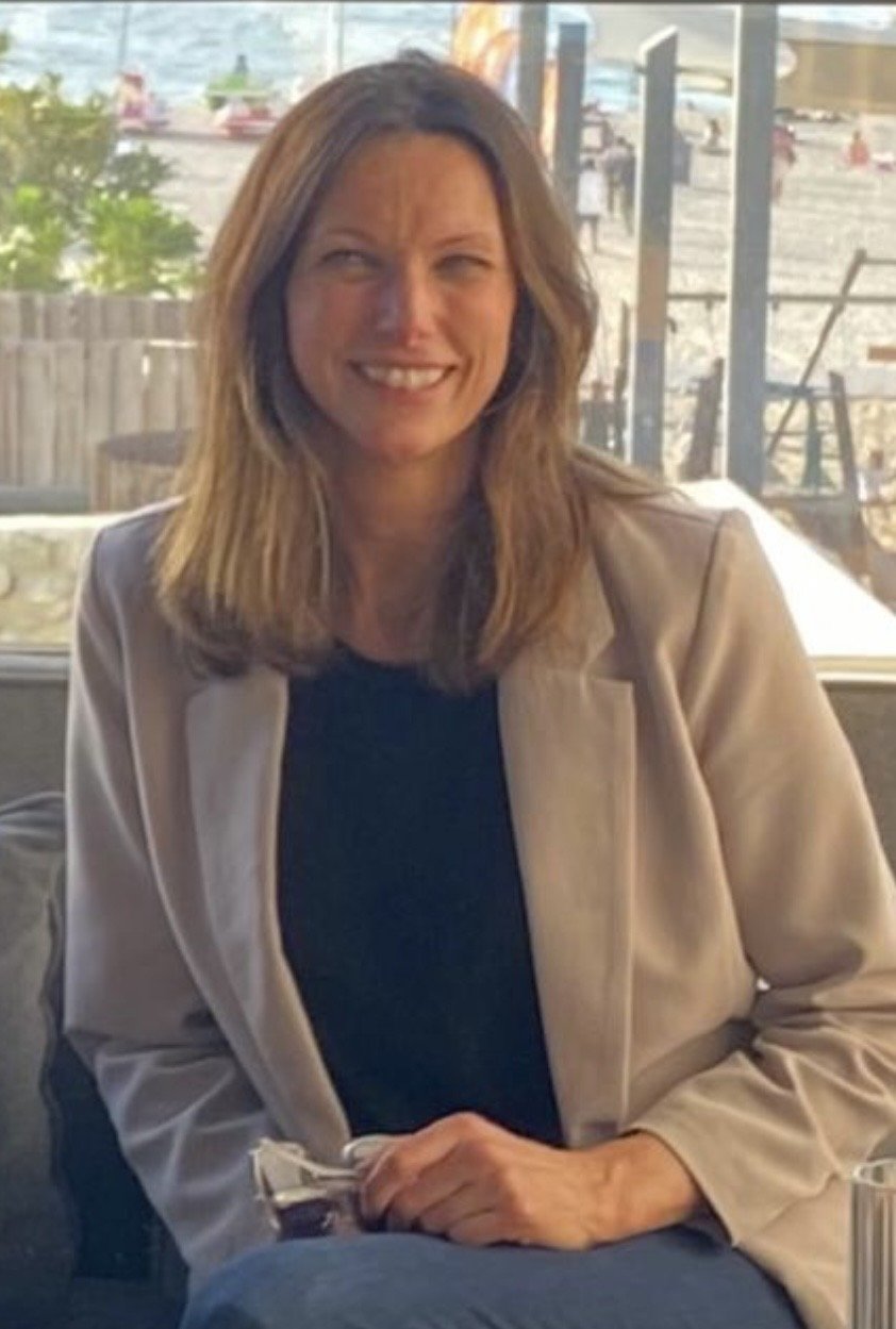 A smiling woman with shoulder-length brown hair wearing a beige jacket and black top, sitting indoors near a large window.