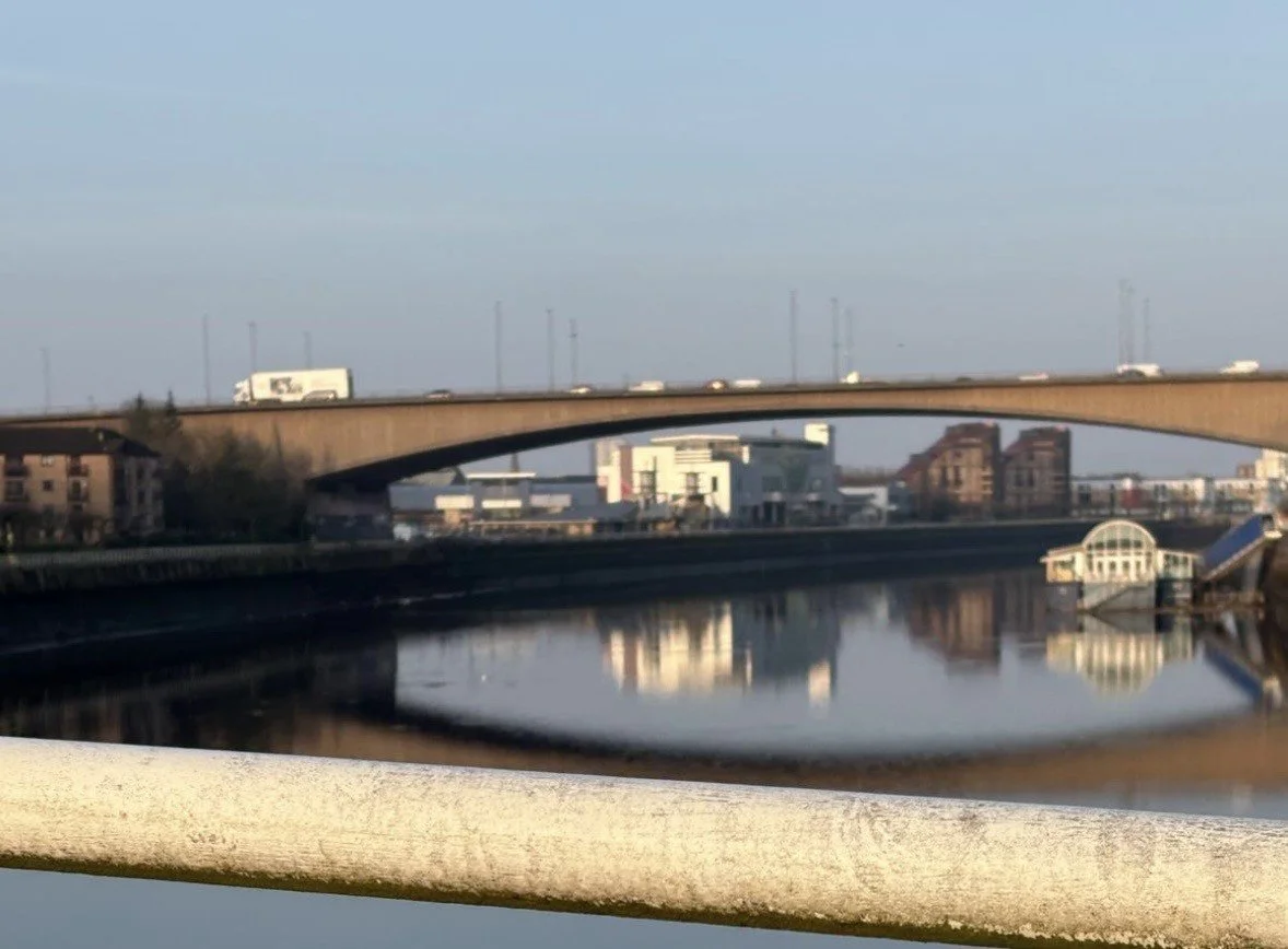 A river with reflections of buildings and bridges, with a bridge showing cars on top and a small building with a rounded roof on the riverbank.