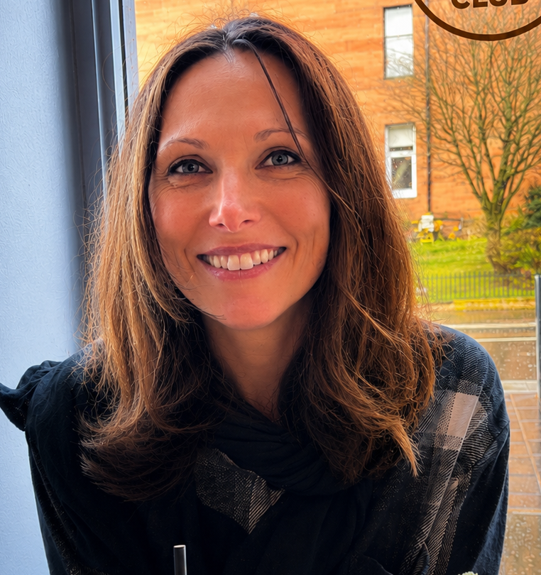 Smiling woman with shoulder-length brown hair sitting indoors near a window, with an outdoor scene of a red brick building and trees in the background.