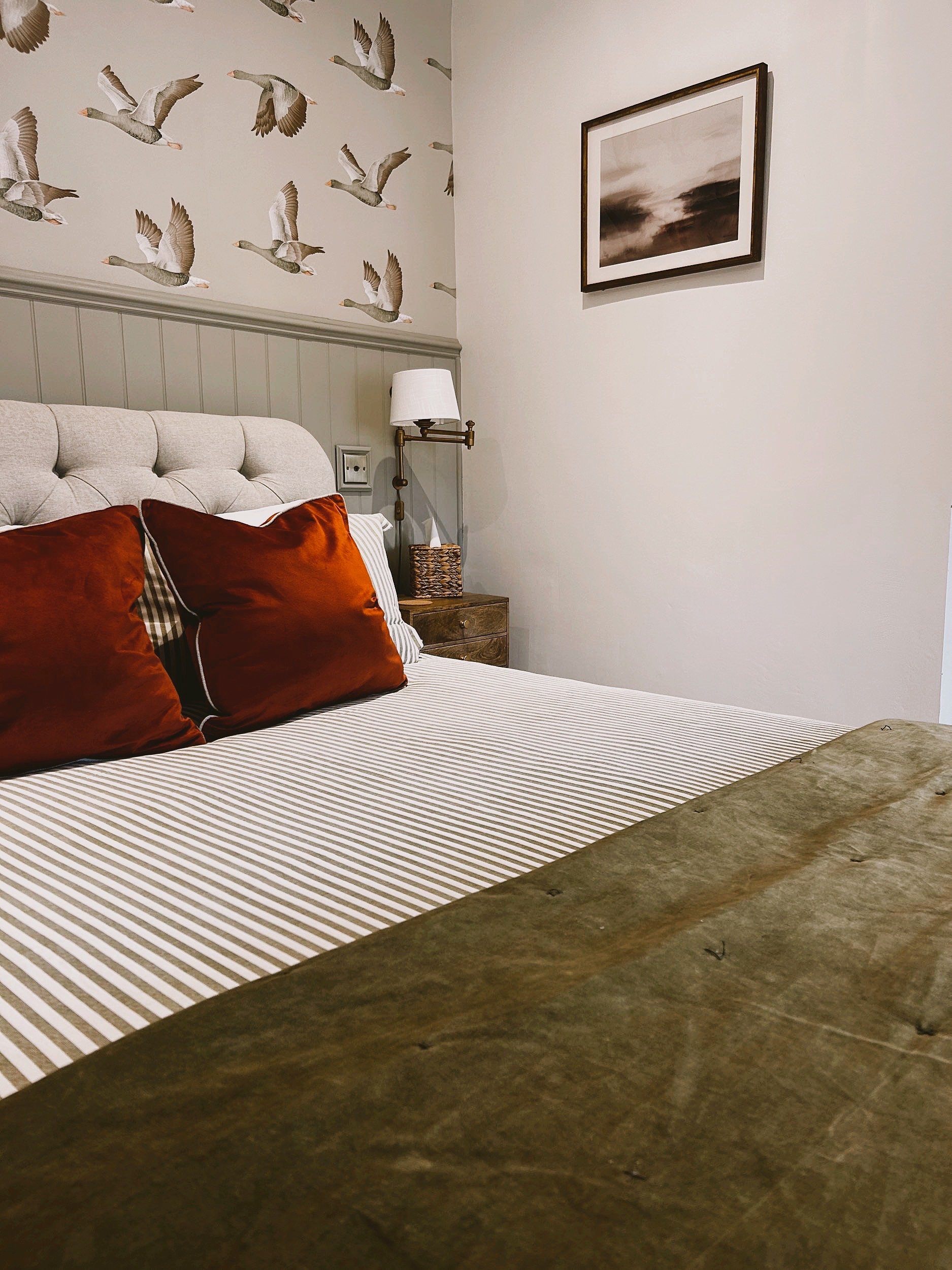 A cozy bedroom with a beige tufted headboard, striped bedding, and rust-colored pillows. A nightstand with a woven basket, white lamp, and a framed artwork of a cloudy sky. Wooden table in the foreground with a striped runner.
