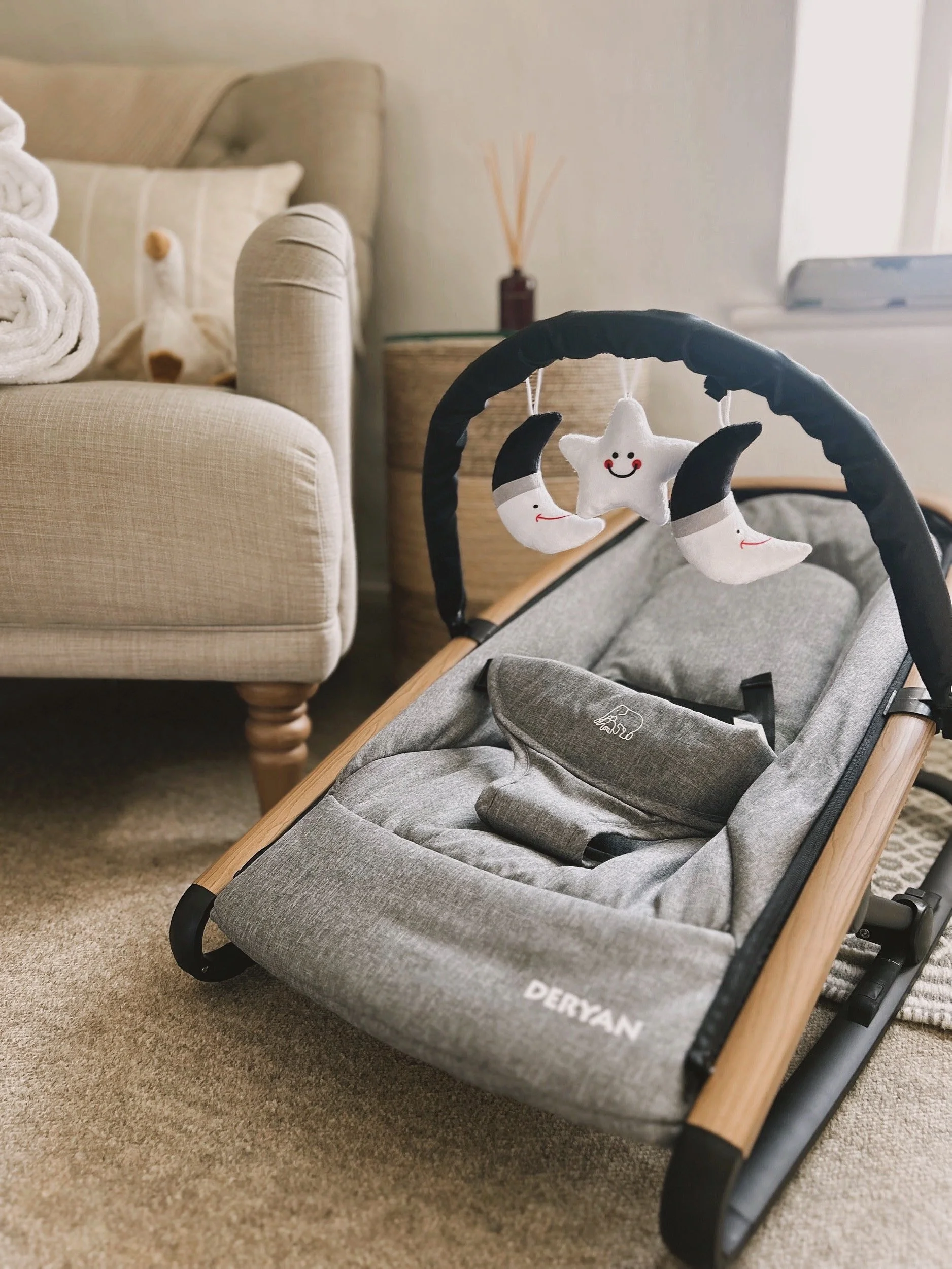 Gray children's stroller with plush moon and star toys hanging from the handle, placed on a beige carpet in a living room with beige sofa and side table.