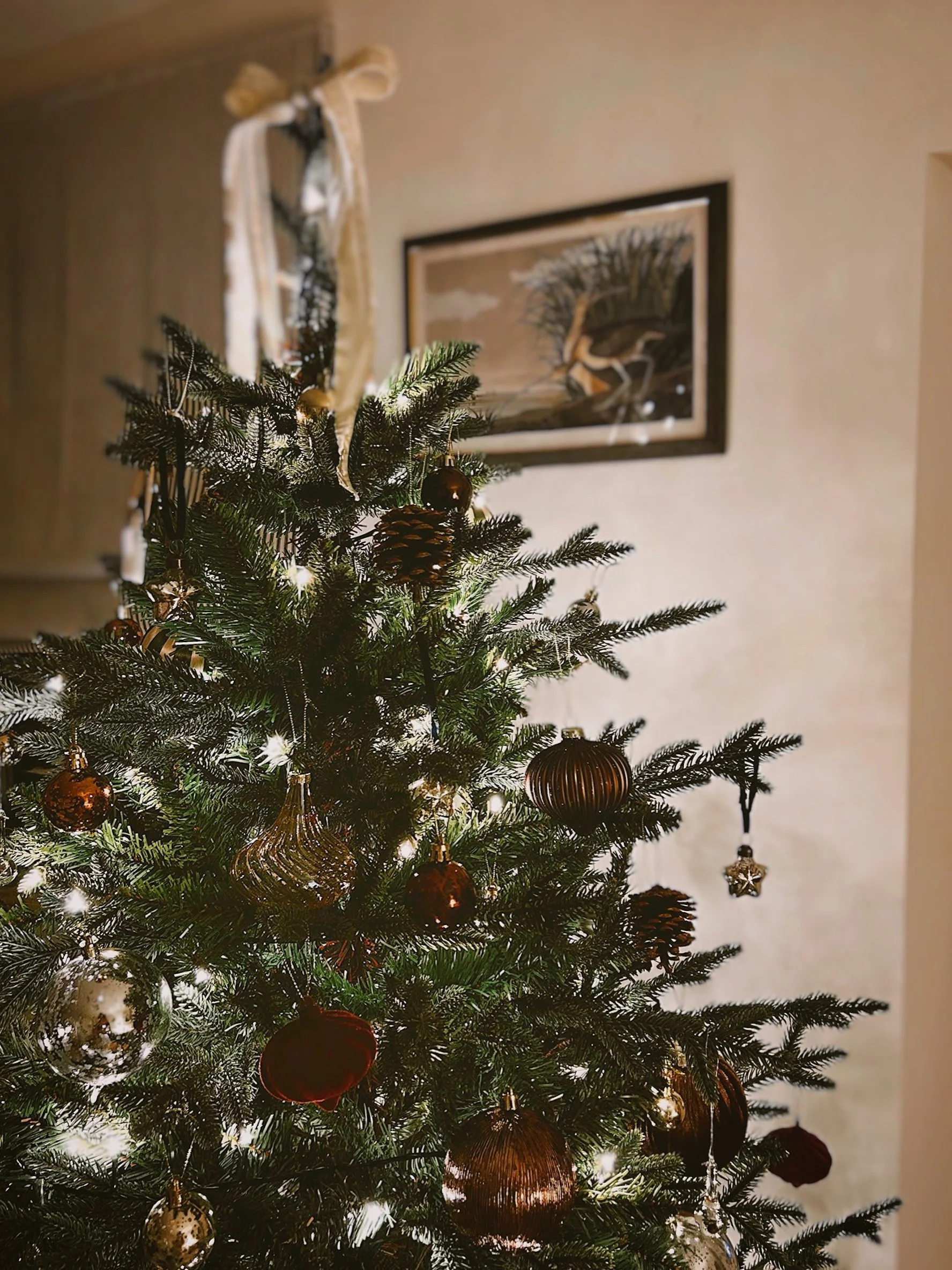 Cottage Christmas tree with glass baubles and pinecones topped with a bow and a bird art piece in the background