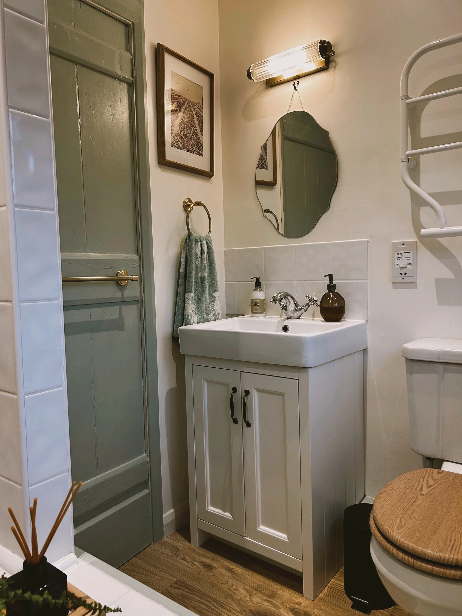 Small bathroom with a white vanity, sink, round mirror, towel rack, and wall-mounted light above. Wooden toilet seat visible next to the sink. Framed artwork and soap dispensers on the counter. Neutral color palette.