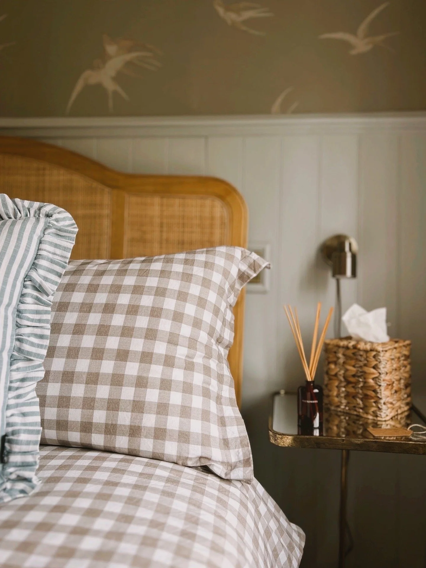 Close-up of a bedroom with a checkered pillow, a wooden headboard, and a bedside table with a reed diffuser and tissue box in a wicker basket.