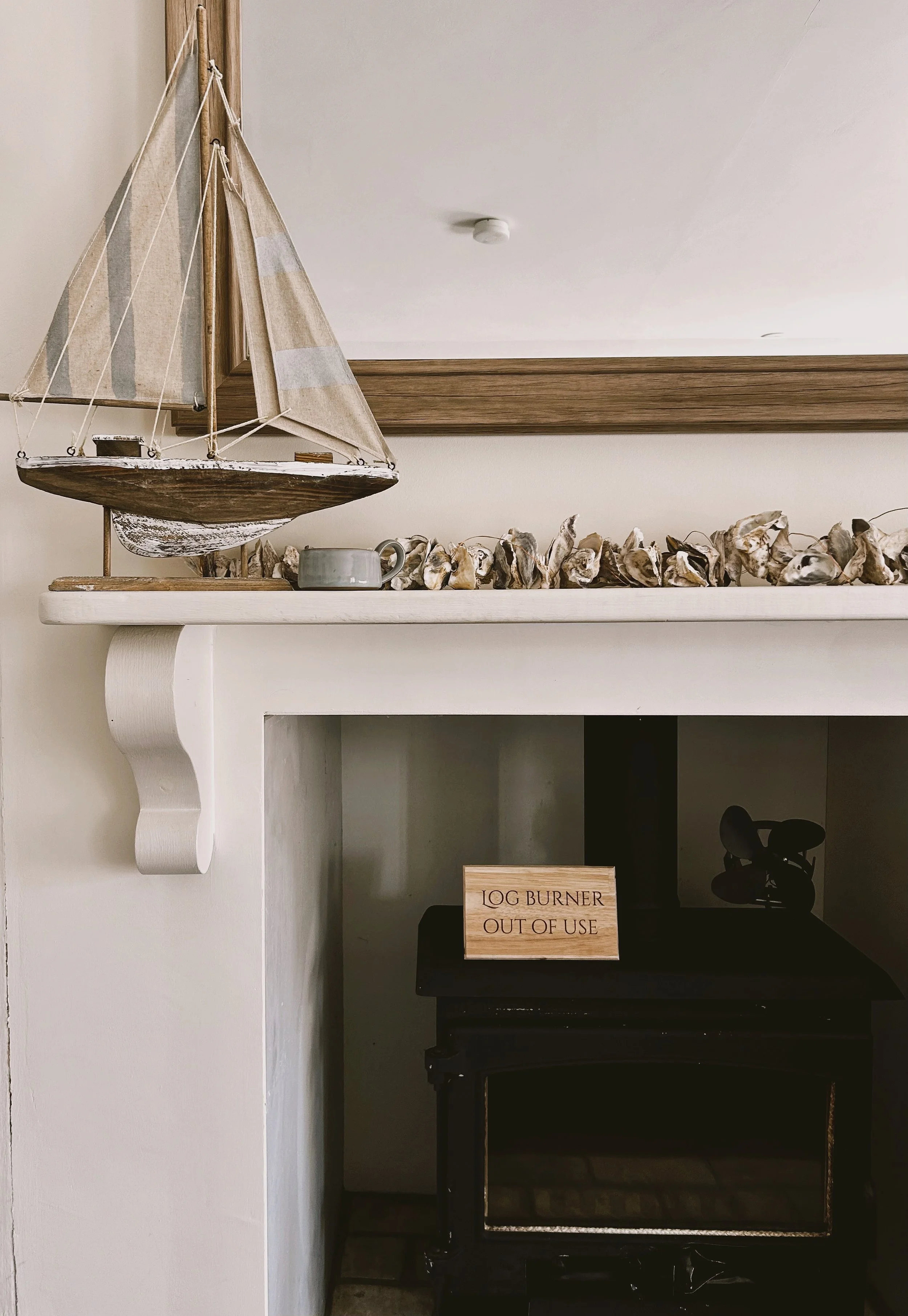 A display shelf holding a model sailboat, a small gray mug, and a row of seashells. Below the shelf is a piece of wood with the text 'LOG BURNER OUT OF USE' and a black stove or fireplace.