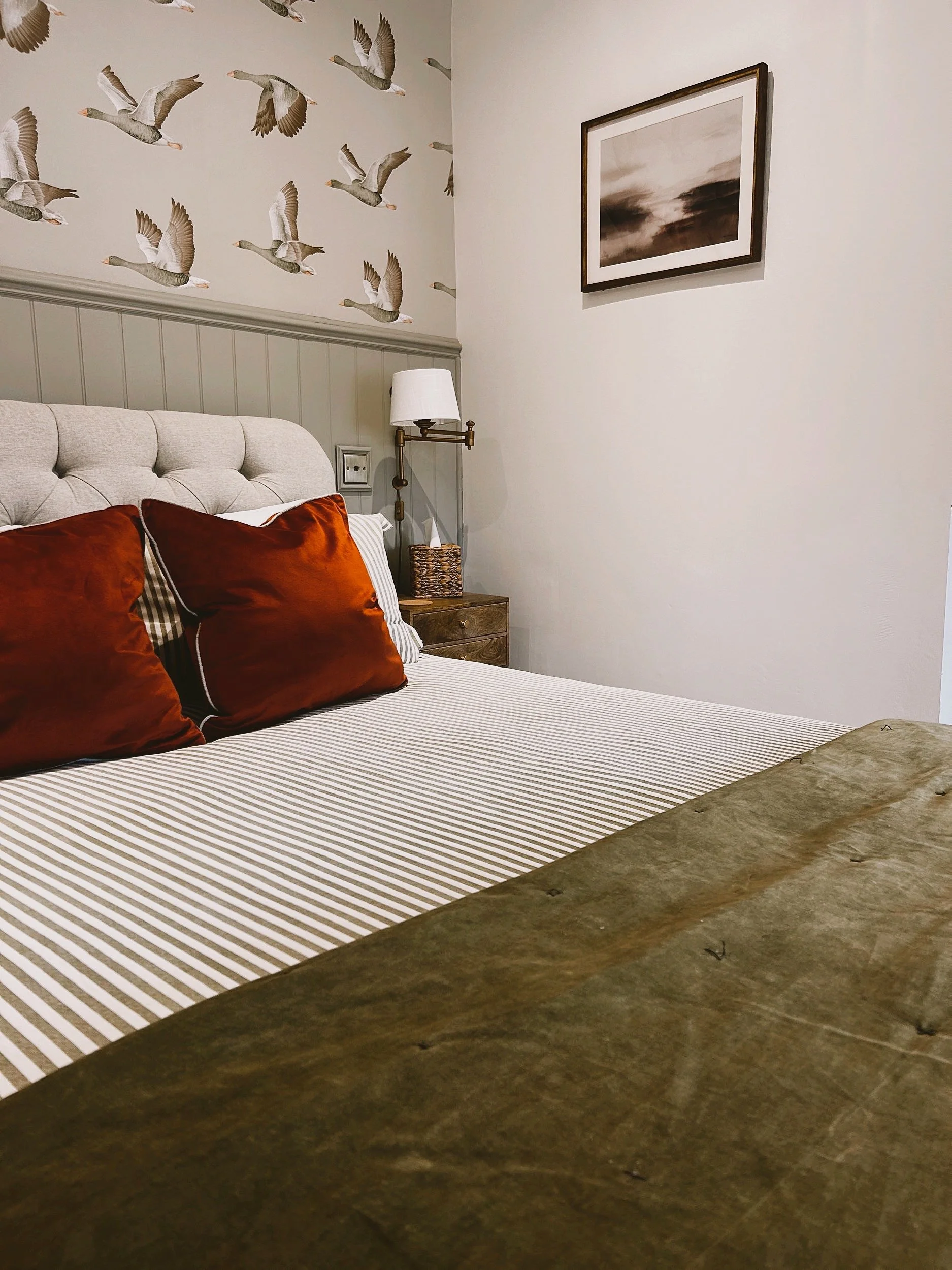 A bedroom with a bed featuring red pillows, a beige tufted headboard, and a wooden nightstand. The wall behind the bed has sander wallpaper with eleysian geese, and there's a framed picture of clouds hanging on the plain white wall.