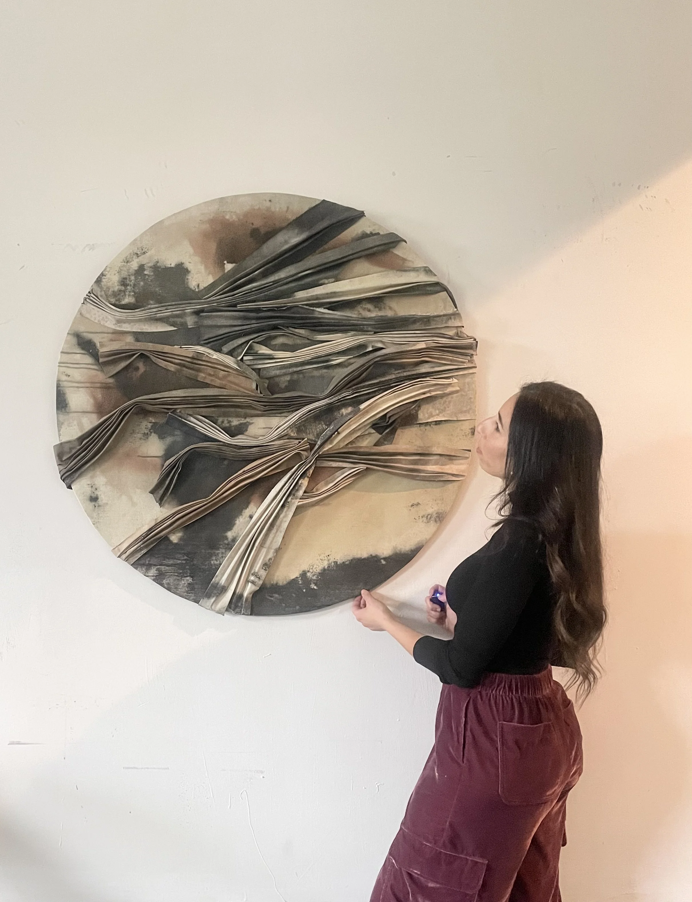 A woman with long dark hair, wearing a black top and maroon pants, stands and looks at a circular abstract art piece on the wall, holding a small object in her right hand.