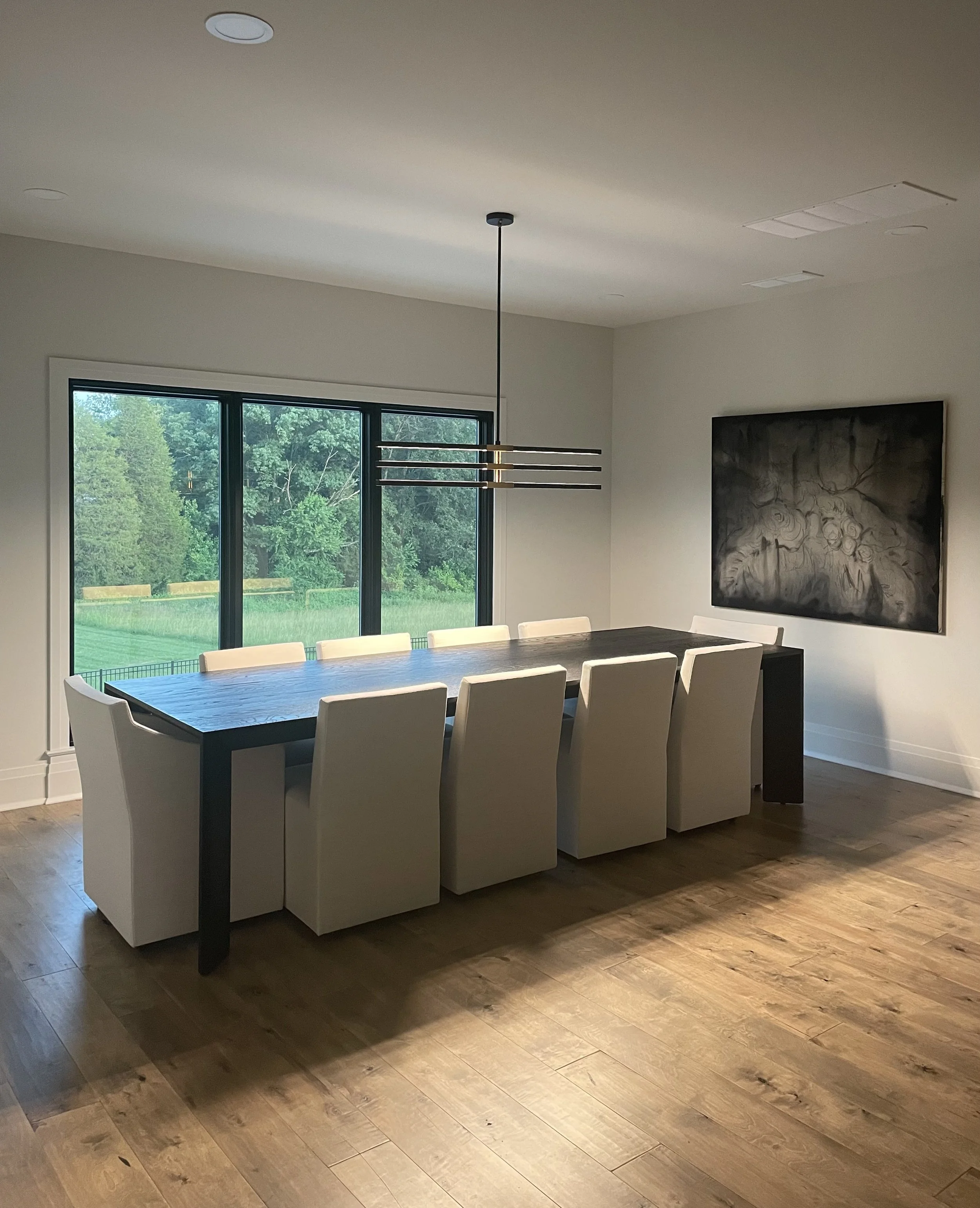 A dining room with a rectangular black table, white chairs with high backs, large windows showing a green outdoor landscape, a black hanging light fixture above the table, a large black-and-white portrait on the wall, and wooden flooring.