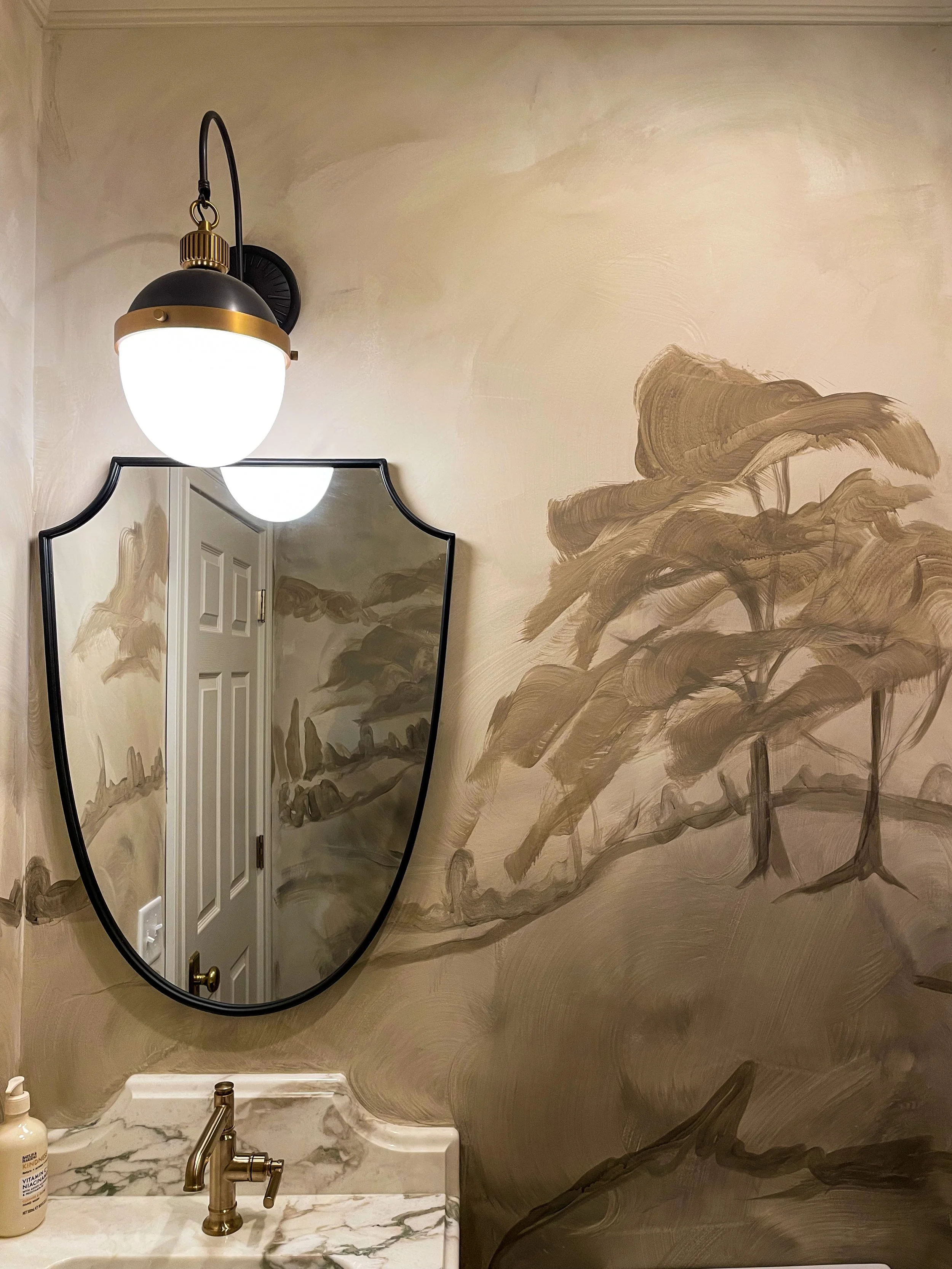 A bathroom wall with painted brownish brushstrokes resembling plants or trees, a mirror reflecting a white door, a black and gold wall sconce with a globe-shaped light, and a marble sink with a gold faucet.