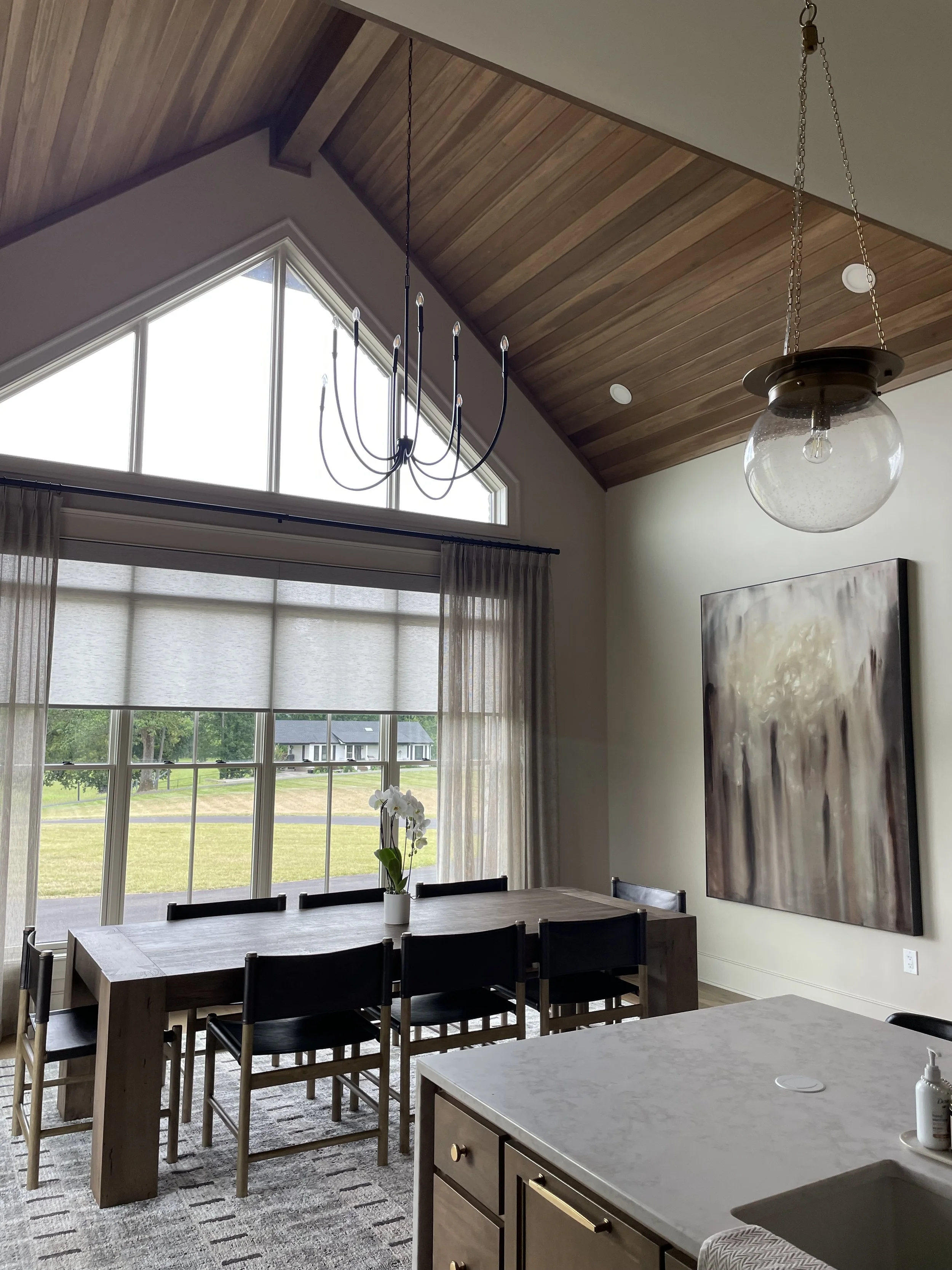 Interior view of a dining area with a wooden table and chairs, a large window with curtains, a modern chandelier, and a pendant light, with a wooden ceiling and art piece on the wall.