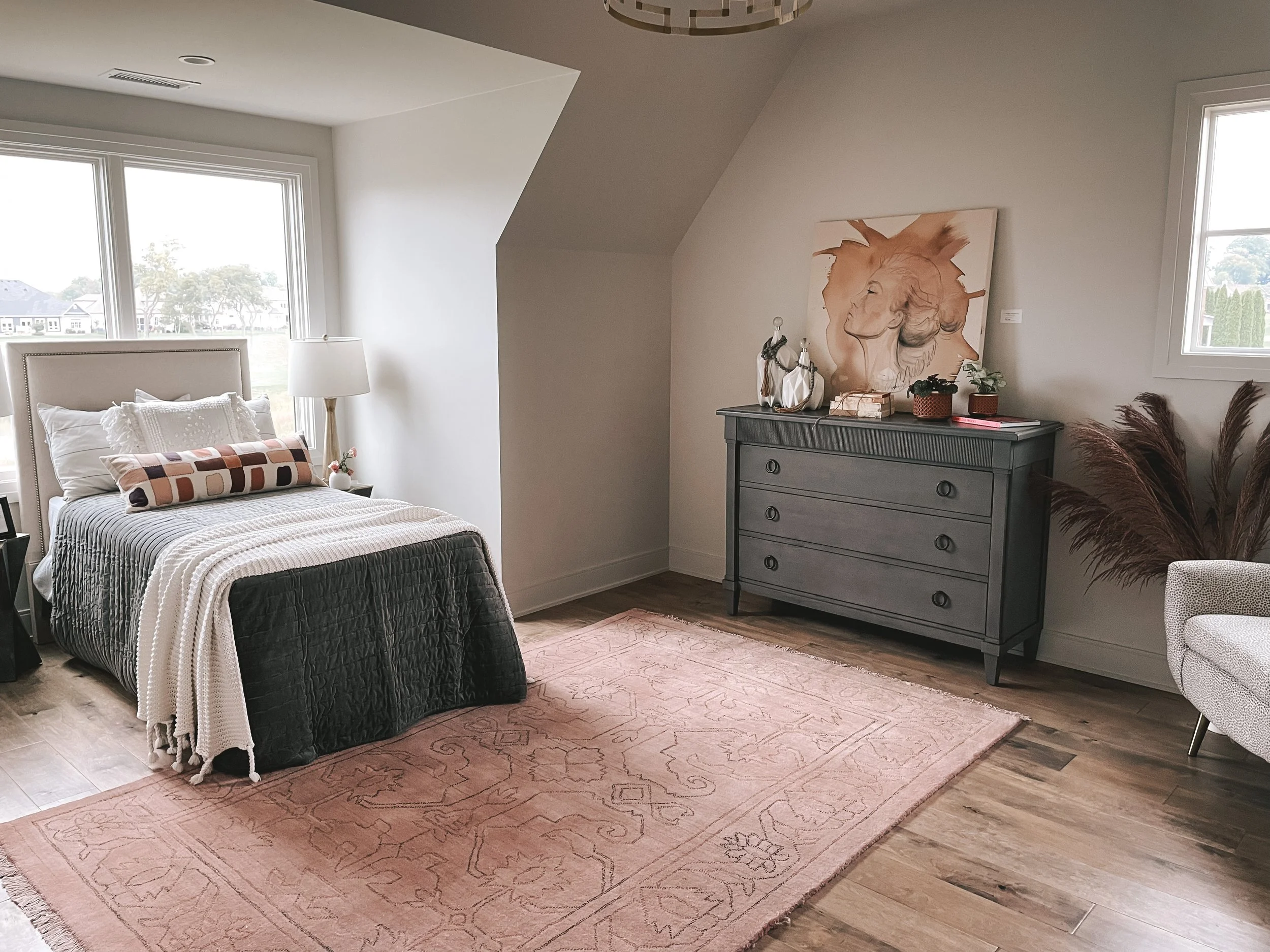 A cozy bedroom with a bed, white headboard, white pillows, and a patterned blanket. There are large windows with a view of trees and houses outside. A black nightstand with a lamp and decorative items is next to the bed. A large pink area rug covers 