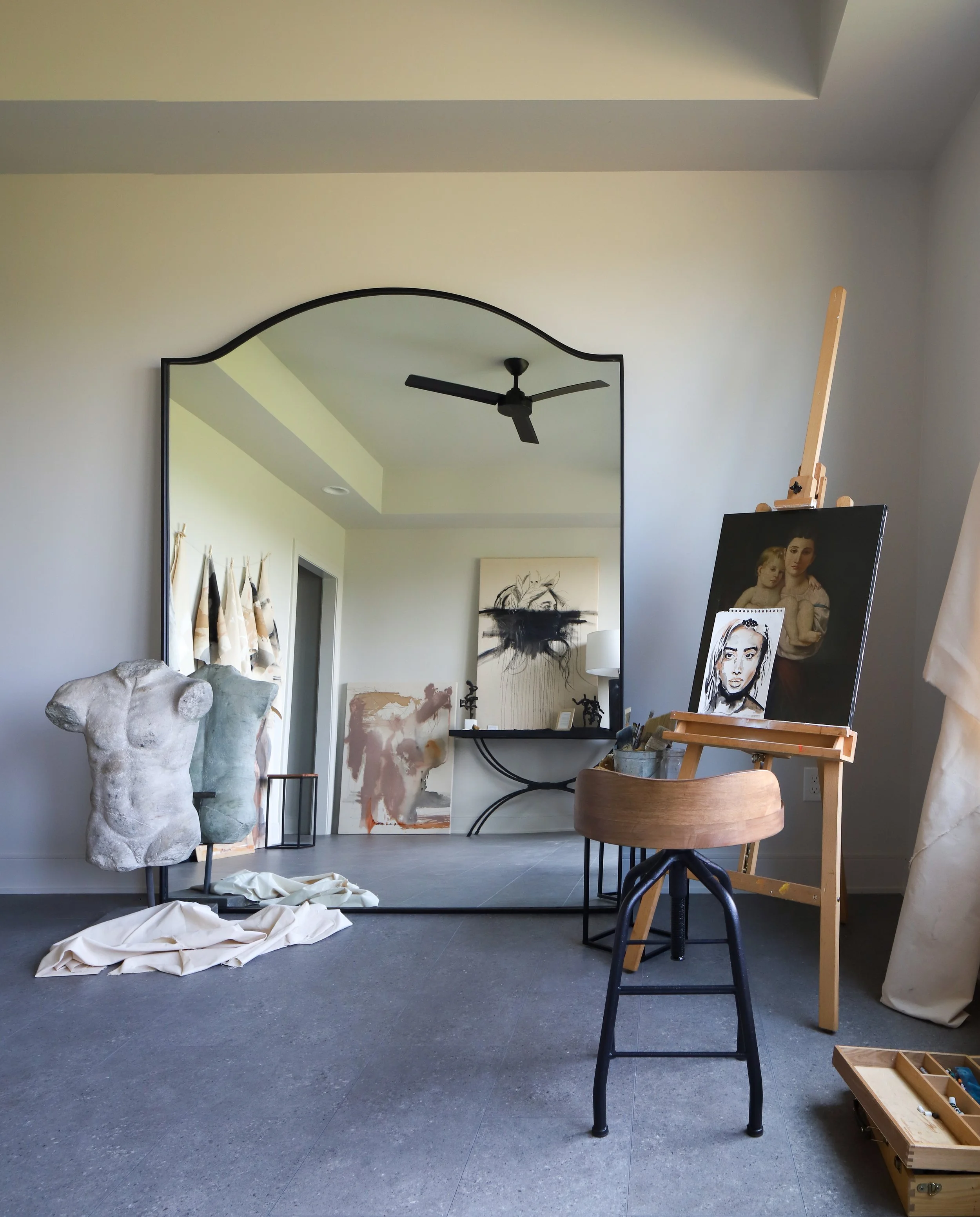 An art studio with a large mirror, sculptures, paintings, and art supplies. A mannequin torso, canvas paintings leaning against the wall, and an easel with artwork. The room has gray flooring and a ceiling fan.