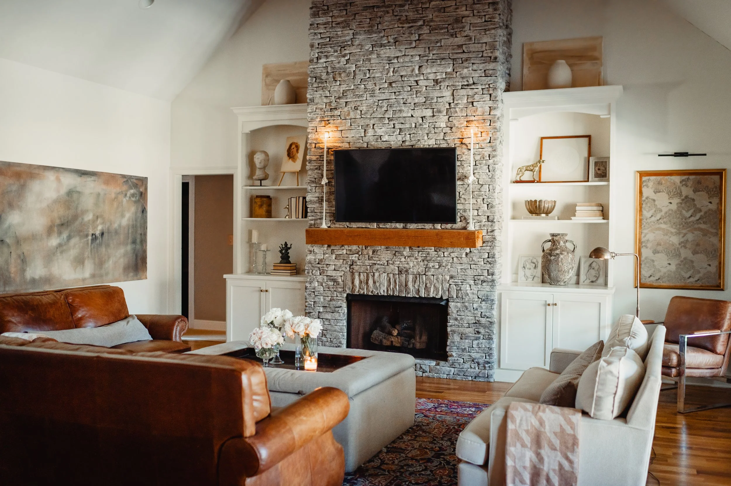 Living room with brick fireplace, television mounted above the fireplace, white built-in shelves with decorative items, brown leather sofas, beige armchair, colorful rug, and floral centerpiece on the coffee table.