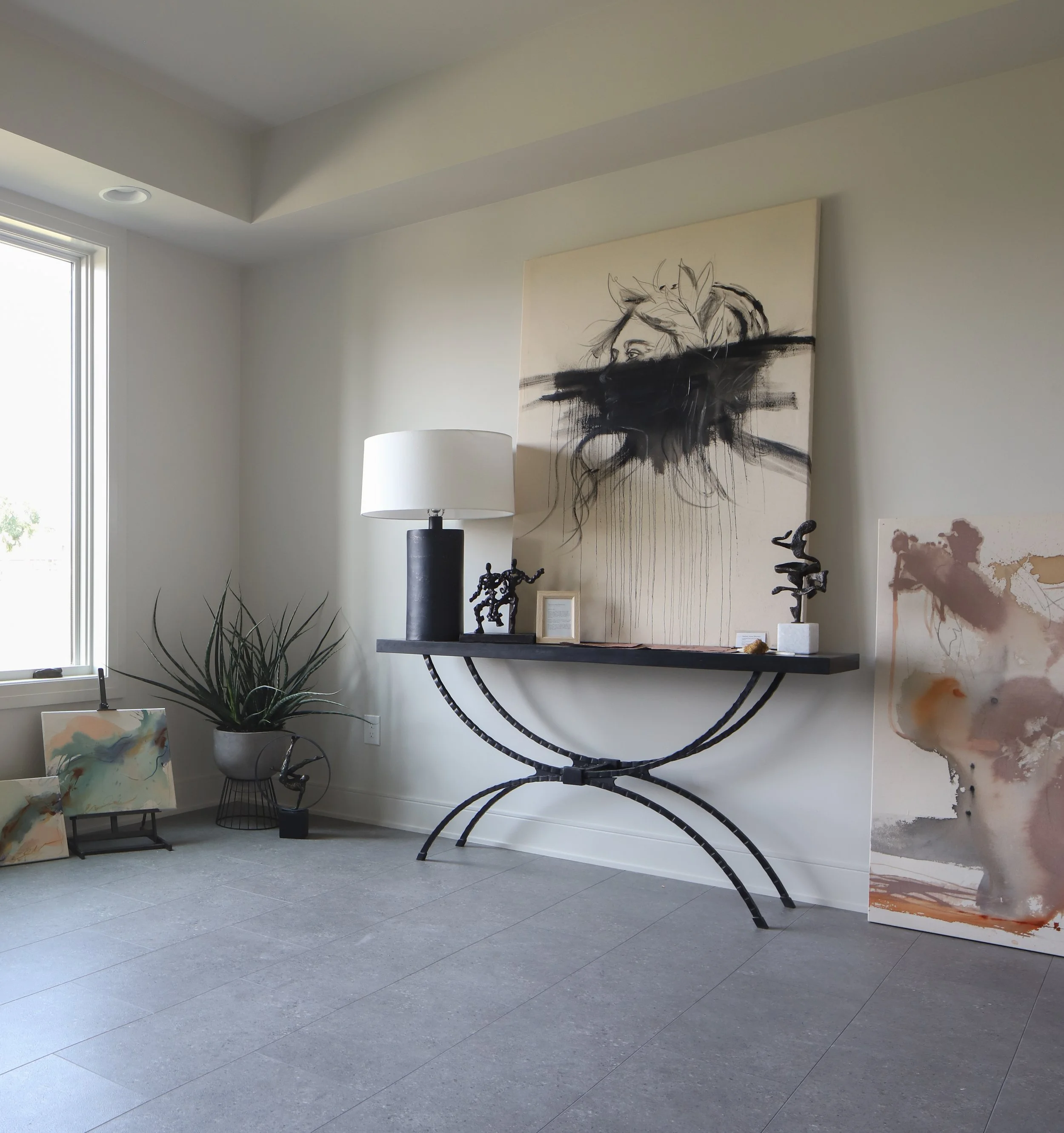Interior view of an art gallery featuring modern artwork and sculptures, with a large window allowing natural light, a white lamp, potted plant, and abstract paintings displayed on the walls and floor.