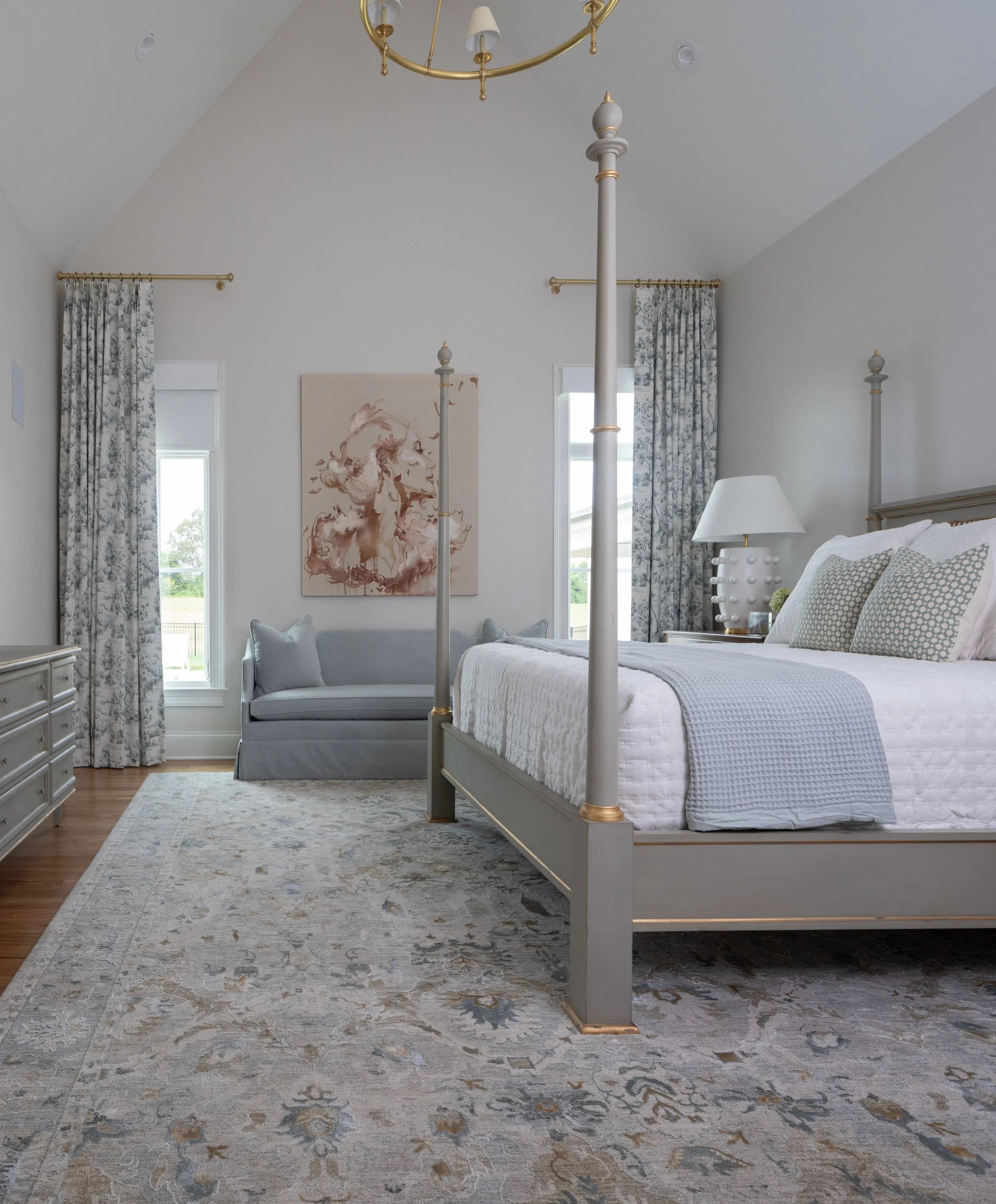 A bedroom with a white canopy bed, a light blue sofa, a white lamp, and floral curtains, with large windows and a decorative painting of a woman on the wall.