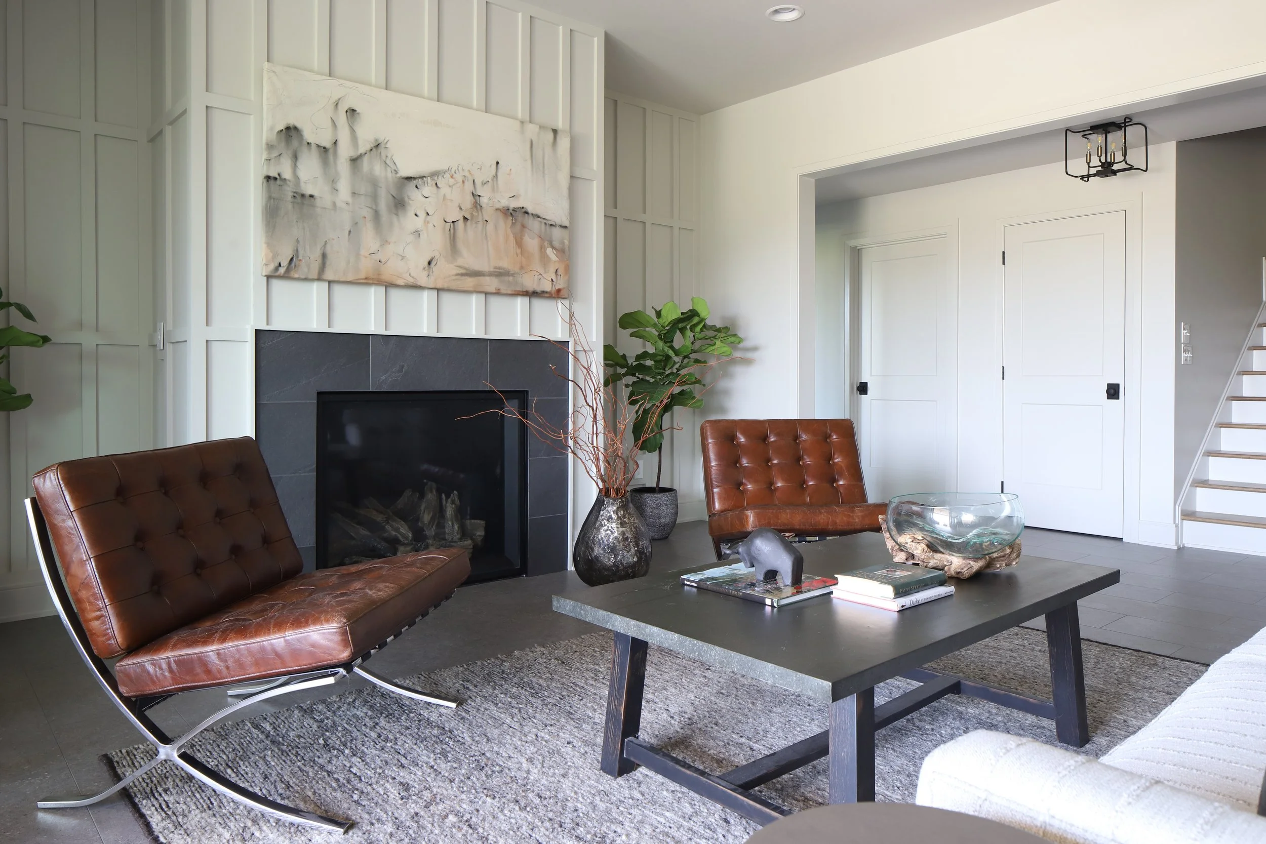 Living room with fireplace, two brown leather chairs, a black coffee table holding books, a decorative glass bowl, and a plant in a black pot. There is a large abstract painting above the fireplace and a modern ceiling light fixture.