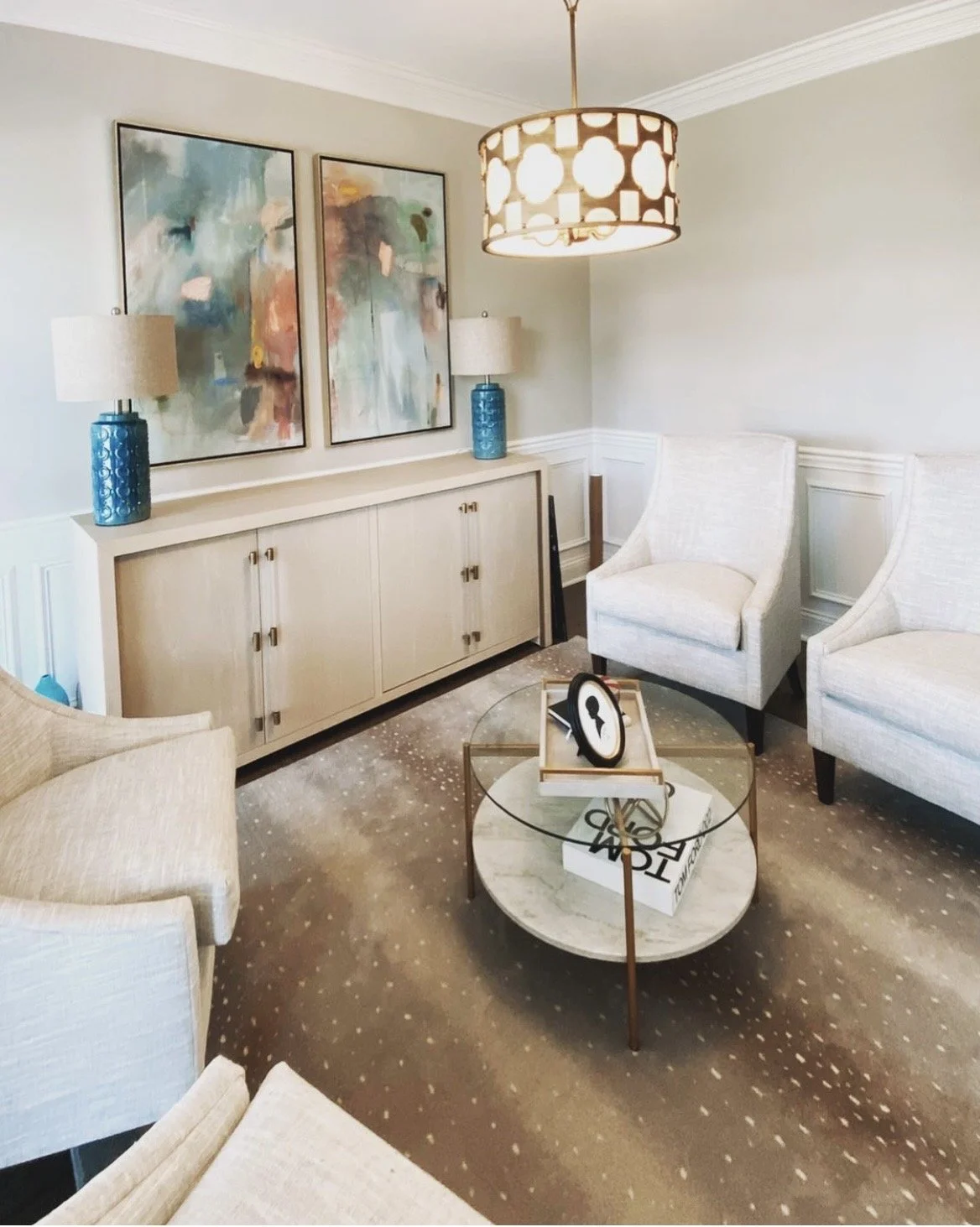 Living room with light-colored walls, a white cabinet with blue lamps, two abstract paintings, white chairs, a glass coffee table, a chandelier, and a patterned beige carpet.
