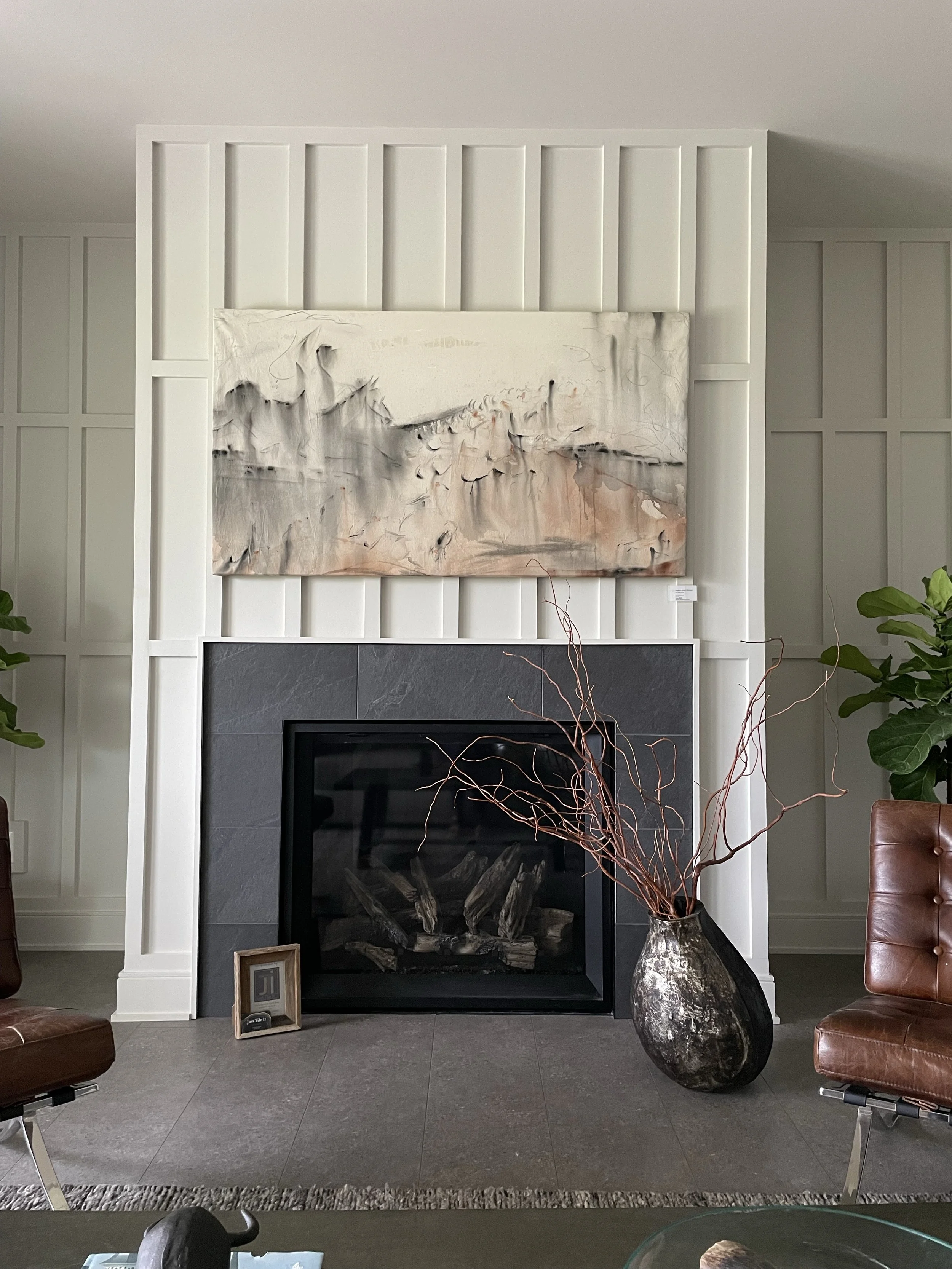 A living room with a modern fireplace framed by white wall paneling, a large abstract painting above the fireplace, a decorative vase with tall, curly branches, and partial views of brown leather chairs. Potted plants are on either side of the firepl