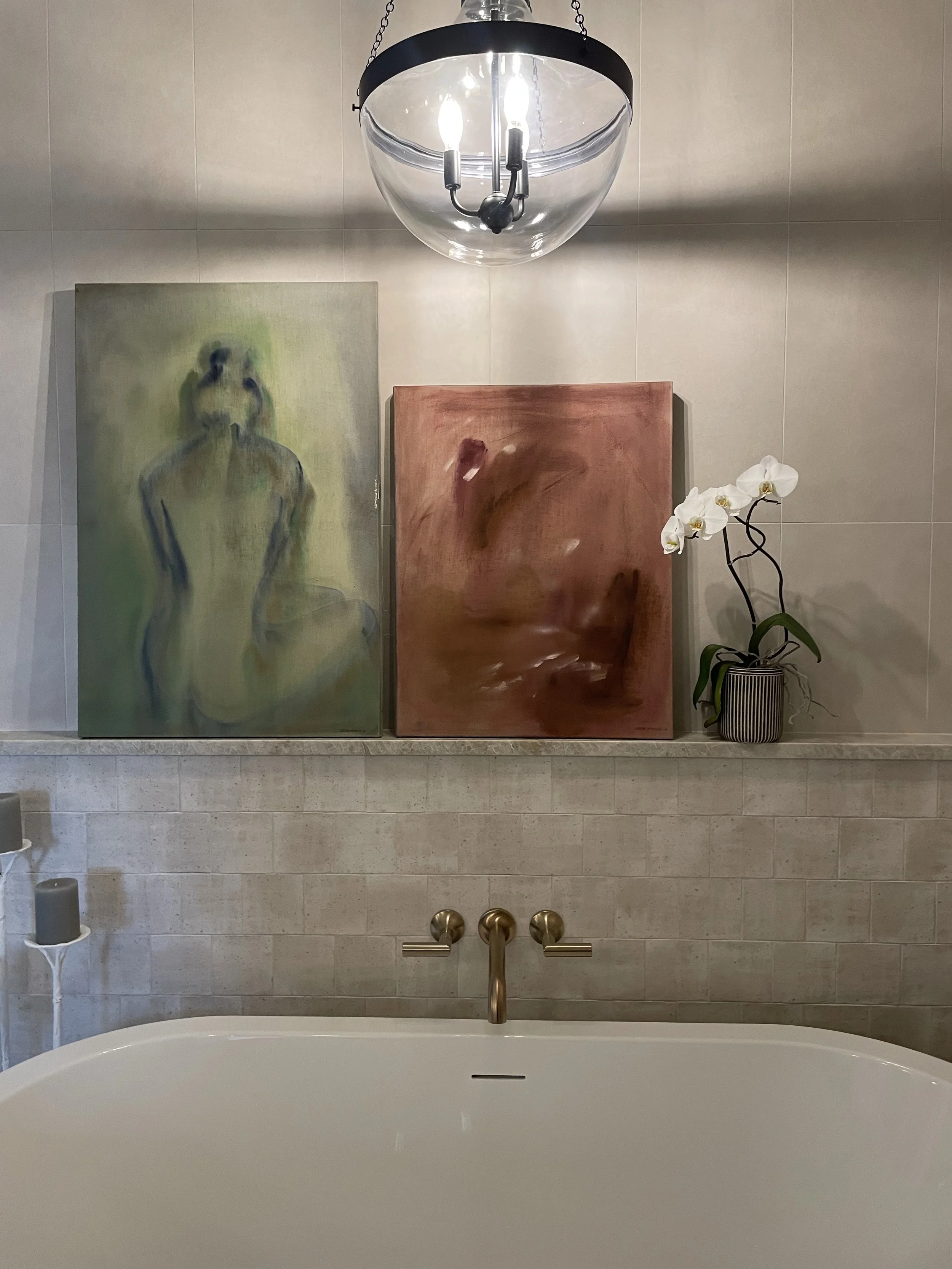 A bathroom mirror reflecting a bathtub faucet, two paintings, and a potted orchid plant, with a chandelier hanging above.