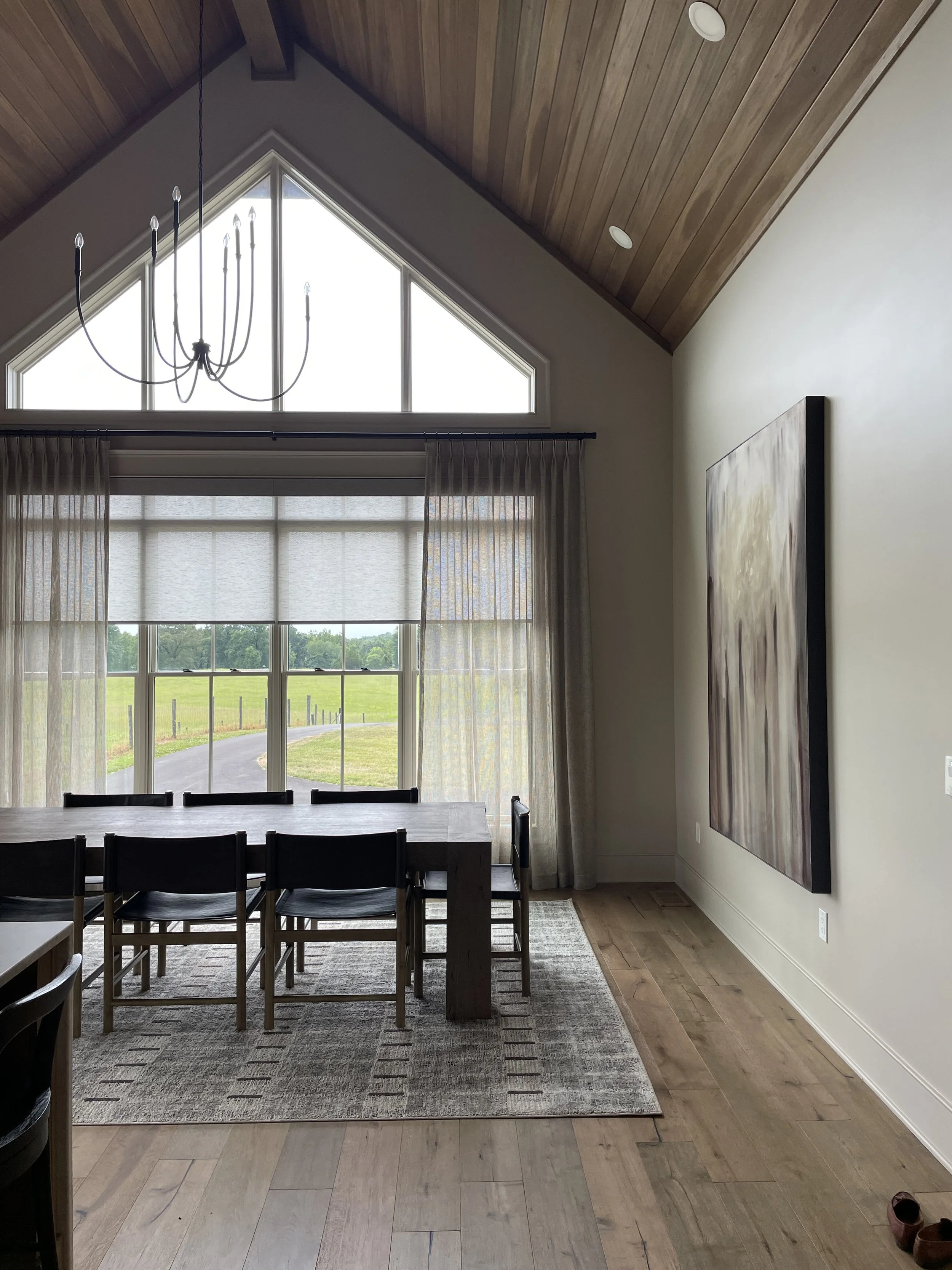 Modern dining area with large windows showing a green outdoor landscape, wooden floor, and ceiling with a chandelier.
