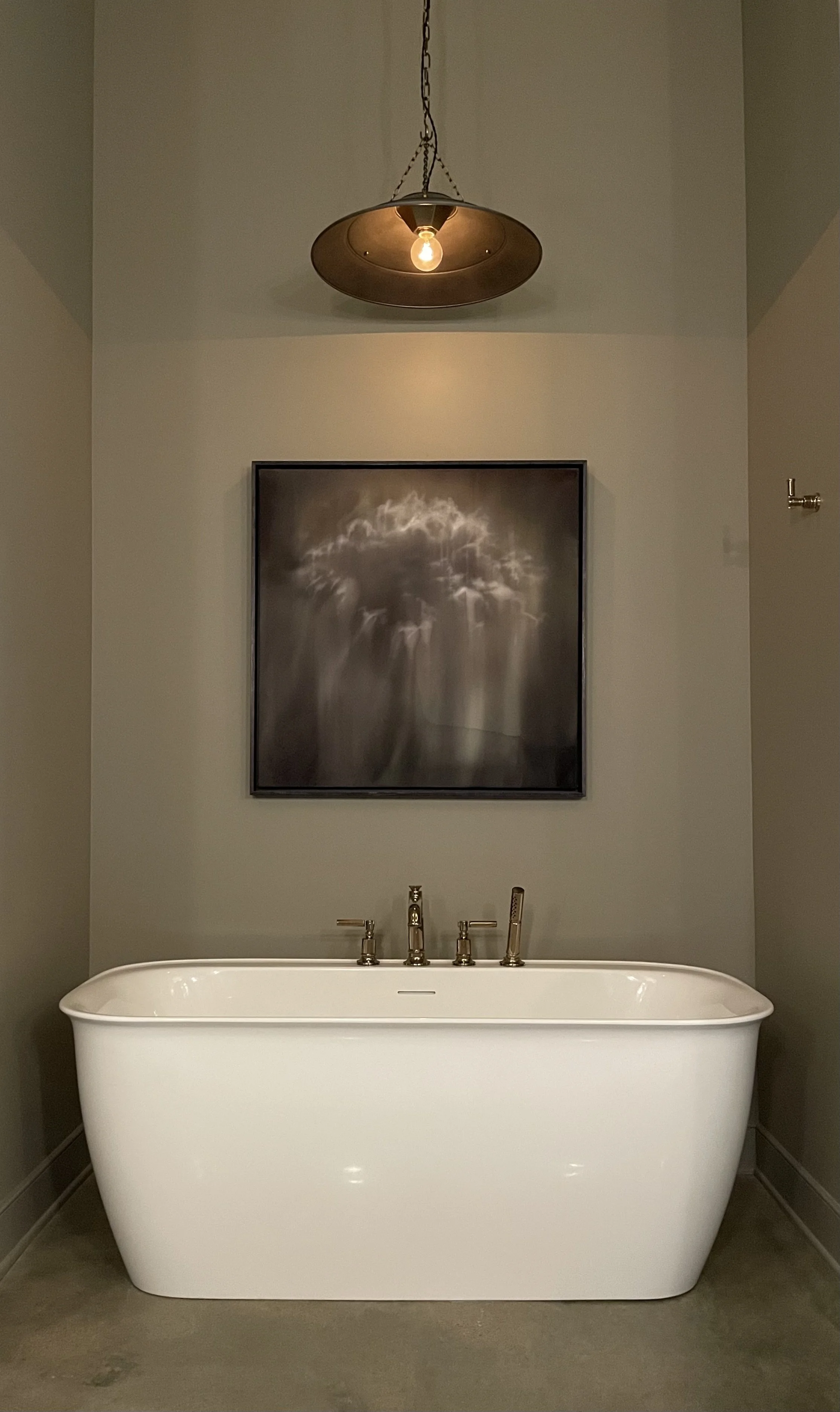 A white freestanding bathtub with silver fixtures in a gray-walled bathroom. Above the tub is a framed black and white photograph of a waterfall, and hanging from the ceiling is a vintage-style pendant light with a single exposed bulb.