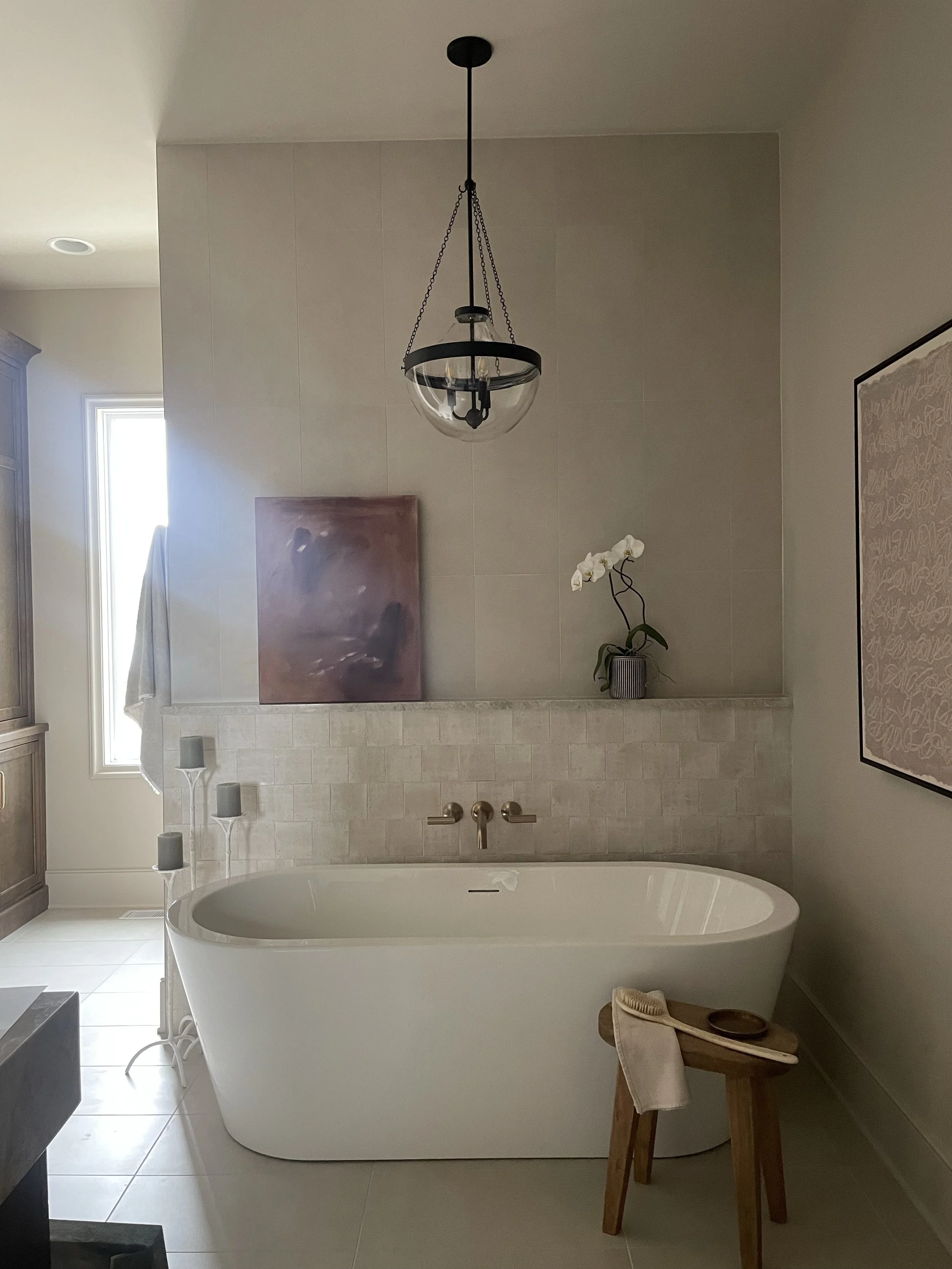 A modern bathroom with a white freestanding bathtub, a small wooden stool with a towel and skin brush, a white orchid in a striped vase, a painting, a chandelier, a window with curtains, and tiled walls and floor.