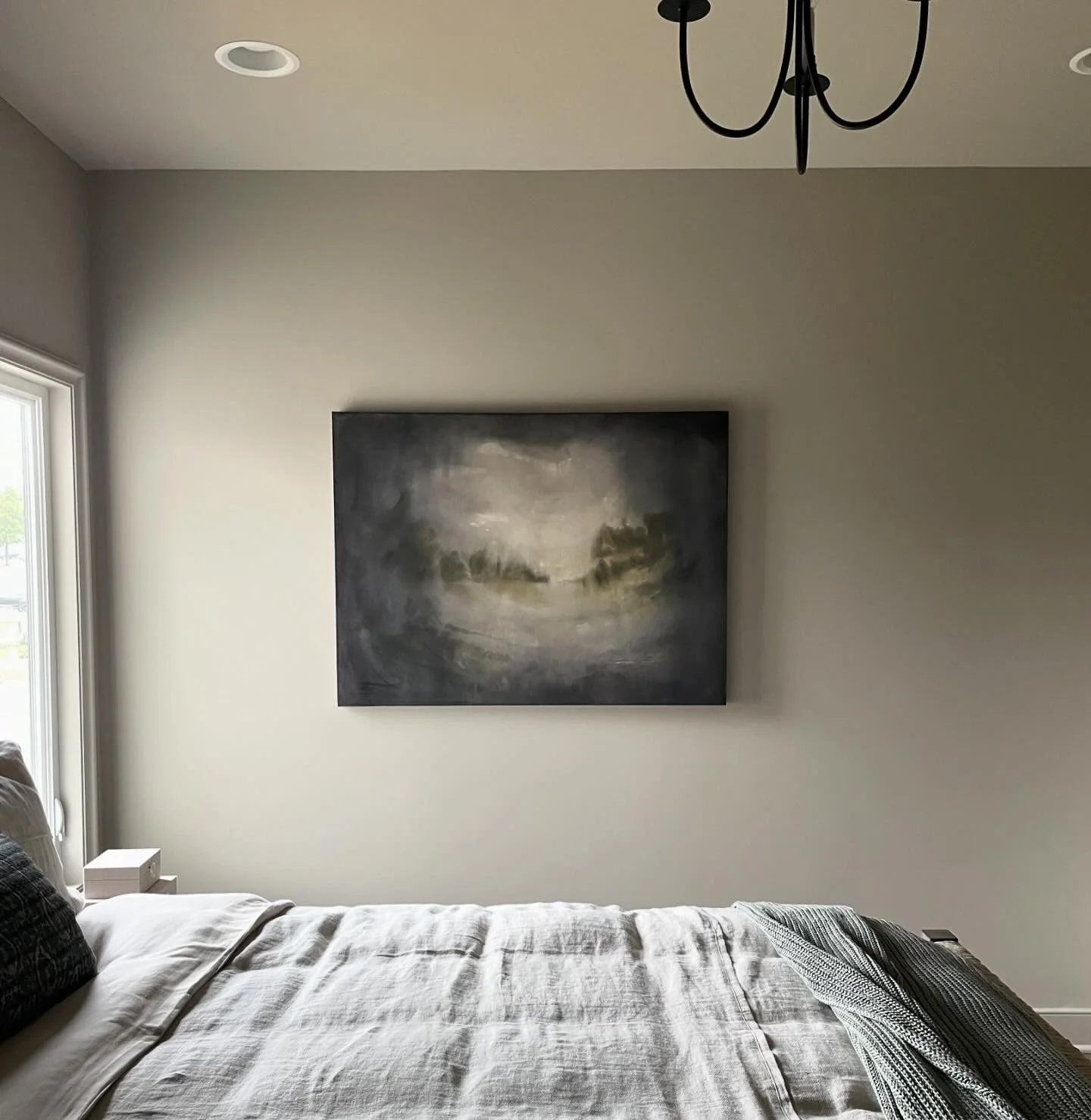A bedroom with a large, abstract landscape painting hanging on a white wall. The bed in the foreground has white and dark pillows, and a striped throw blanket. There is a window on the left, a nightstand with a tissue box, and a ceiling chandelier.