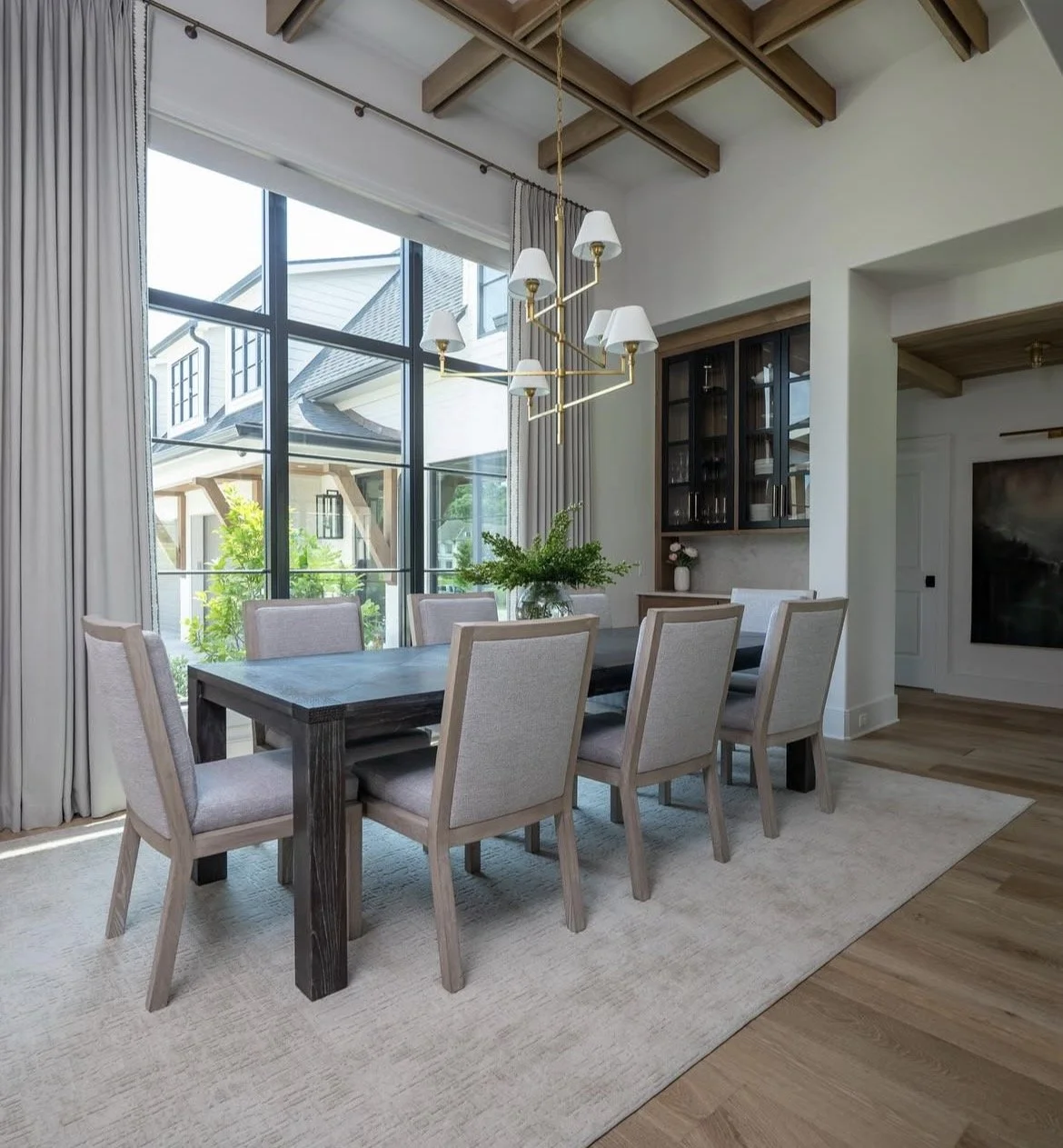 Modern dining room with large windows, a wooden table, beige upholstered chairs, a white rug, a black china cabinet, a green plant, and a gold chandelier.