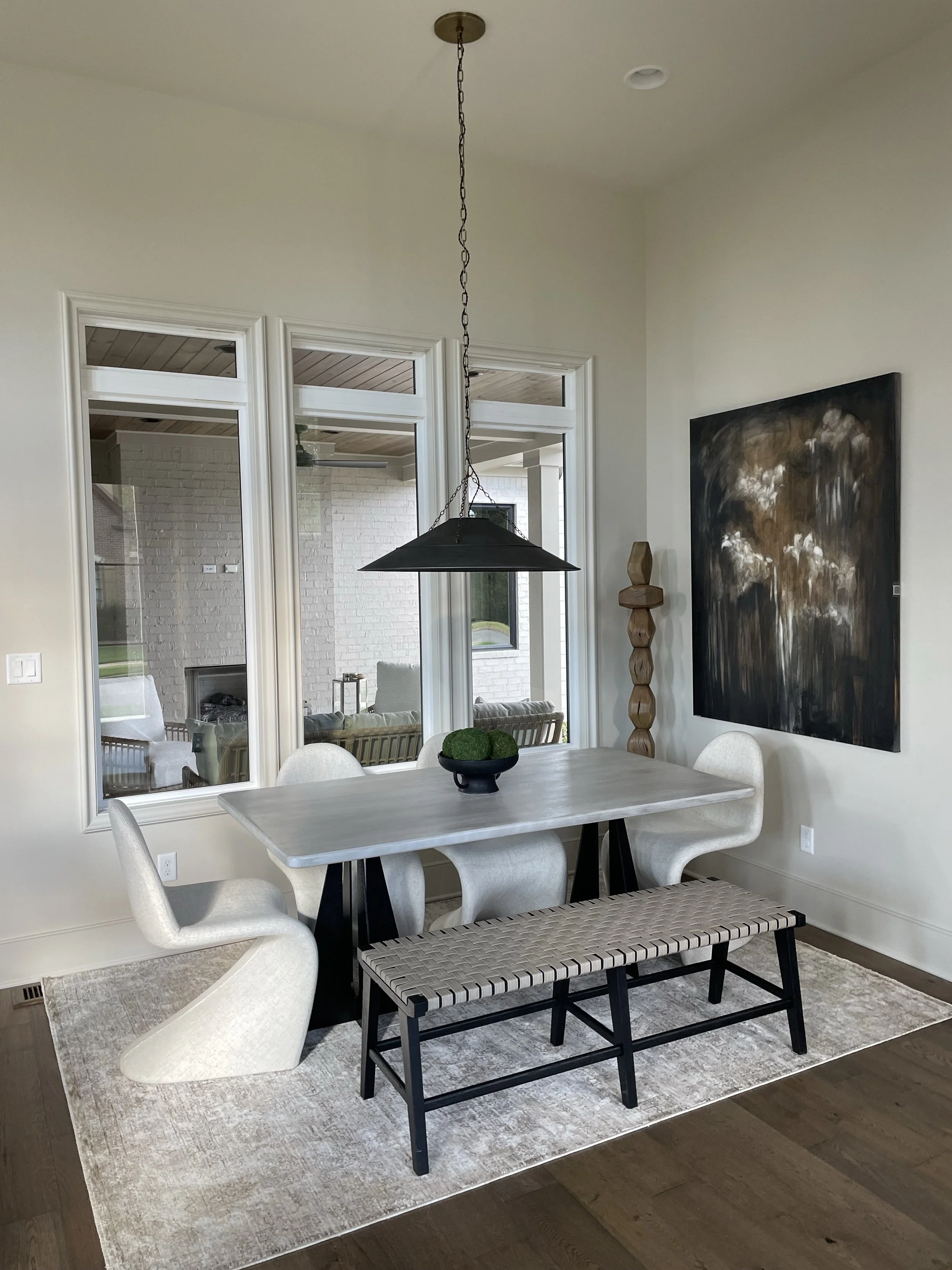 A dining room with a white table, cream-colored modern chairs, a black bench with a woven seat, a large abstract black and brown painting on the wall, a black bowl with green moss centerpiece, and a metal pendant light hanging above the table. The ro
