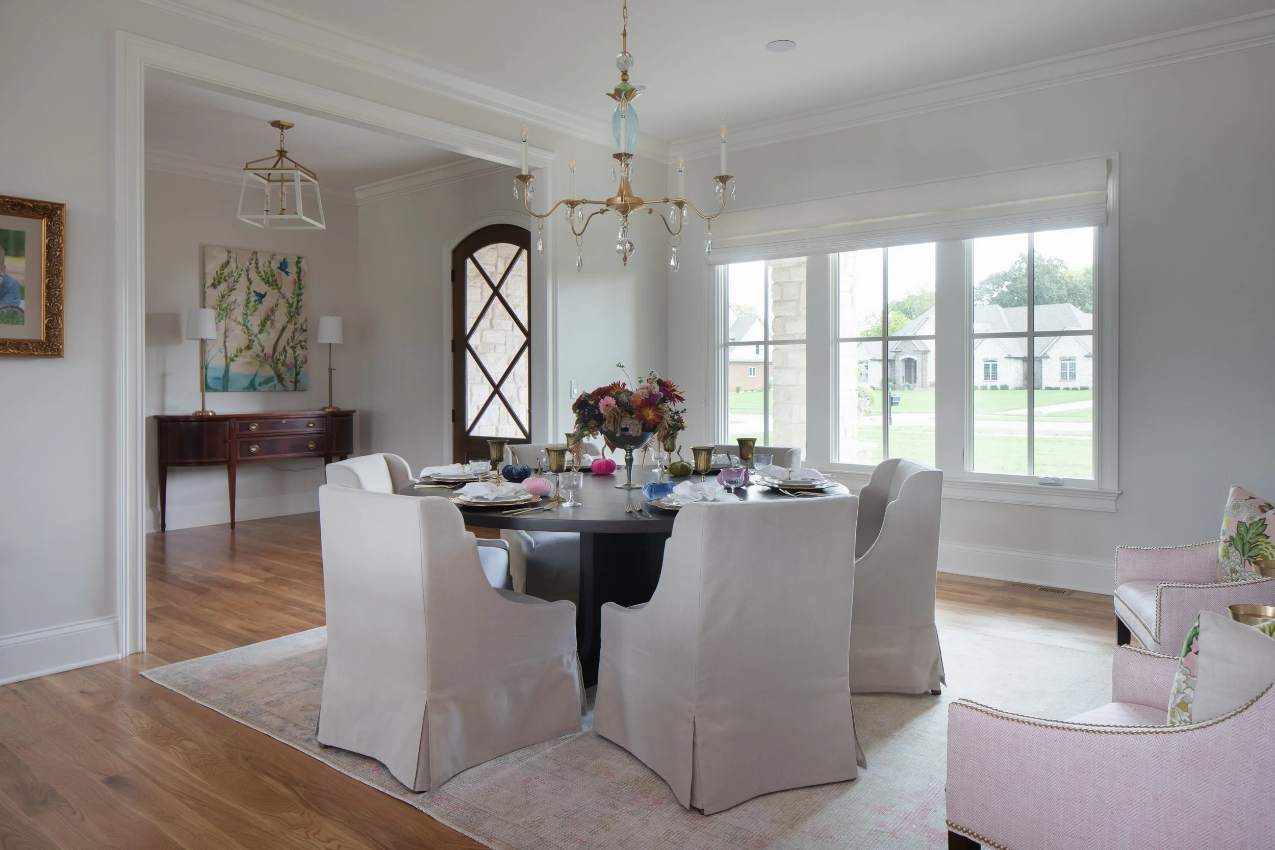 A dining room with a round table set for a meal, surrounded by six white chairs. The table is decorated with a flower arrangement and colorful pumpkins. Large windows allow natural light to fill the room. A chandelier hangs from the ceiling, and a si