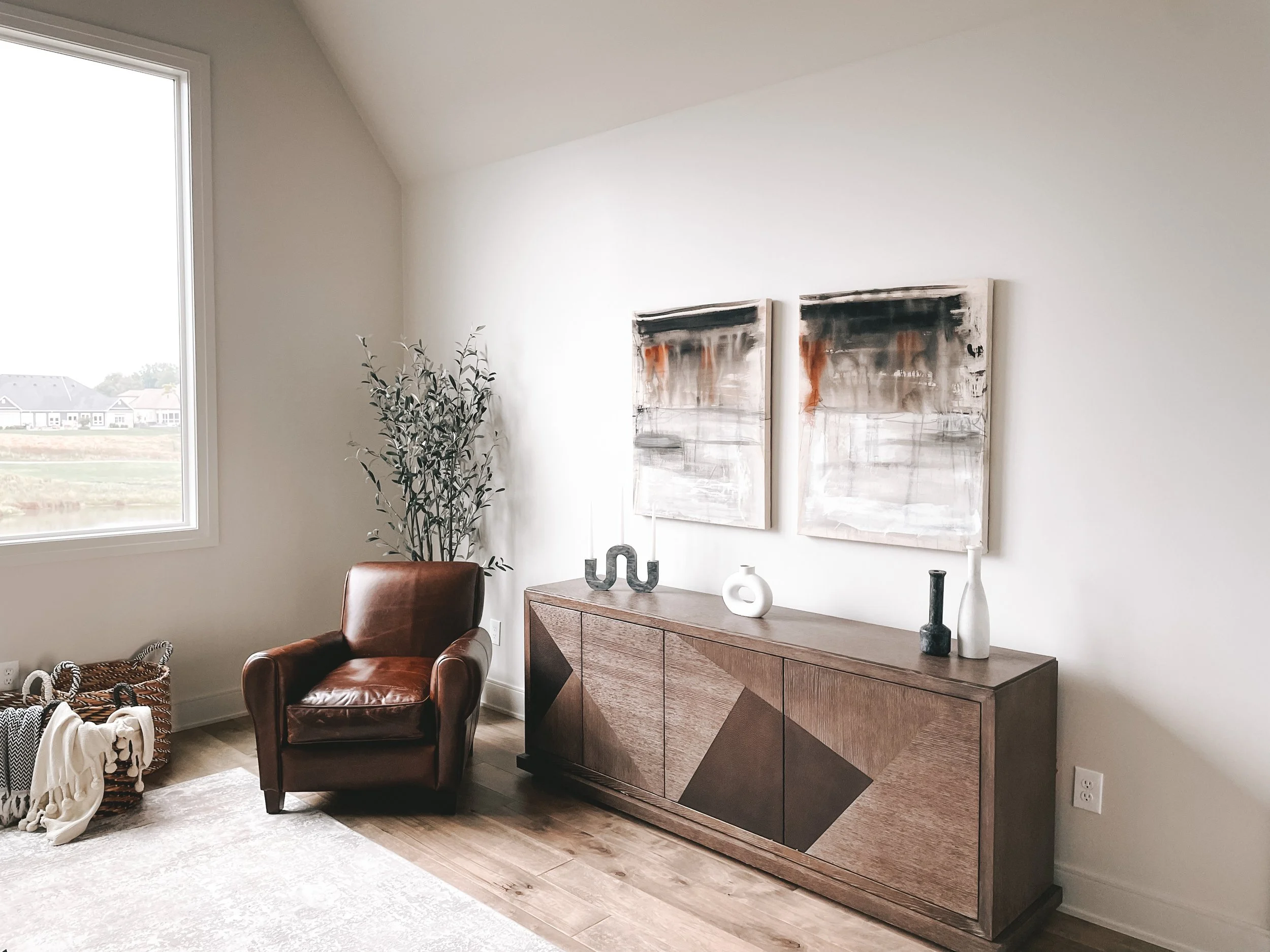 A corner of a living room with a large window, abstract paintings on the wall, a wooden sideboard decorated with vases, a leather chair, and a basket with blankets.