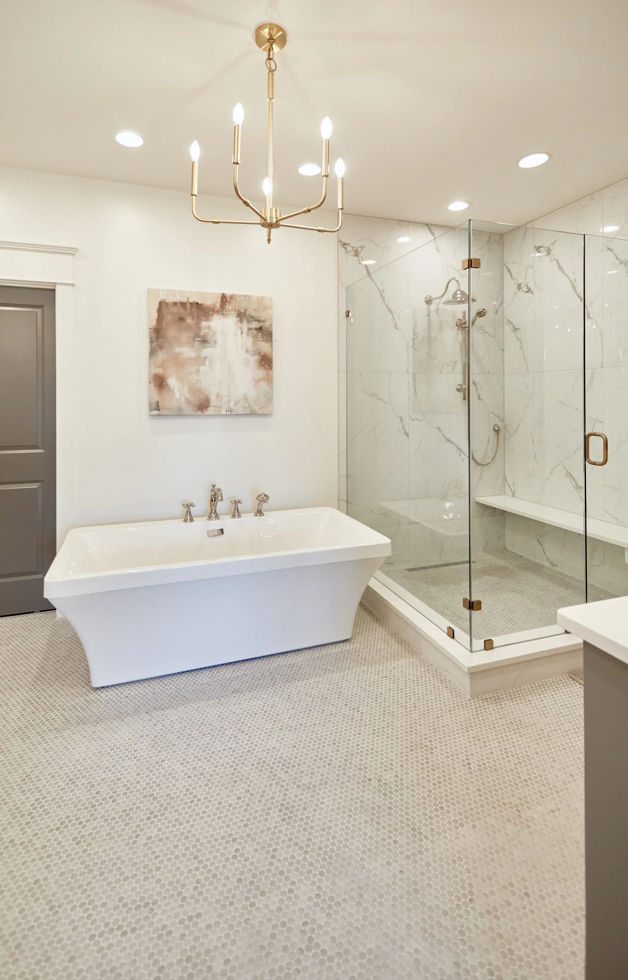 A modern bathroom featuring a white freestanding bathtub, a glass shower enclosure with marble walls, a gold chandelier, and a piece of abstract art on the wall.