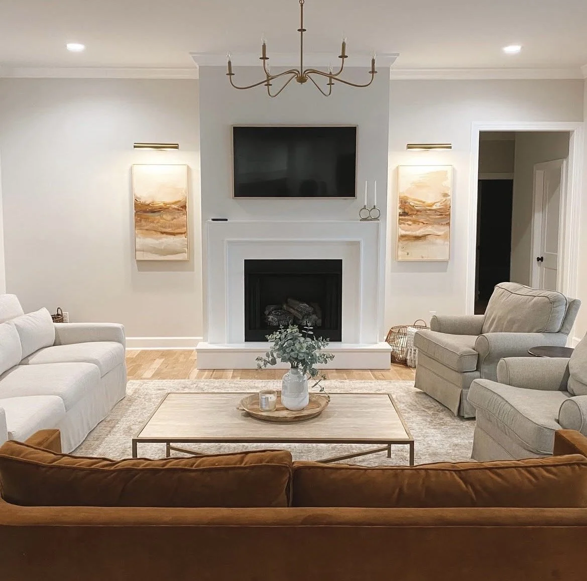 A cozy living room with a white sofa, beige armchairs, and a coffee table with a vase of greenery. There is a fireplace with a TV mounted above it, modern artwork on the walls, and a chandelier hanging from the ceiling.