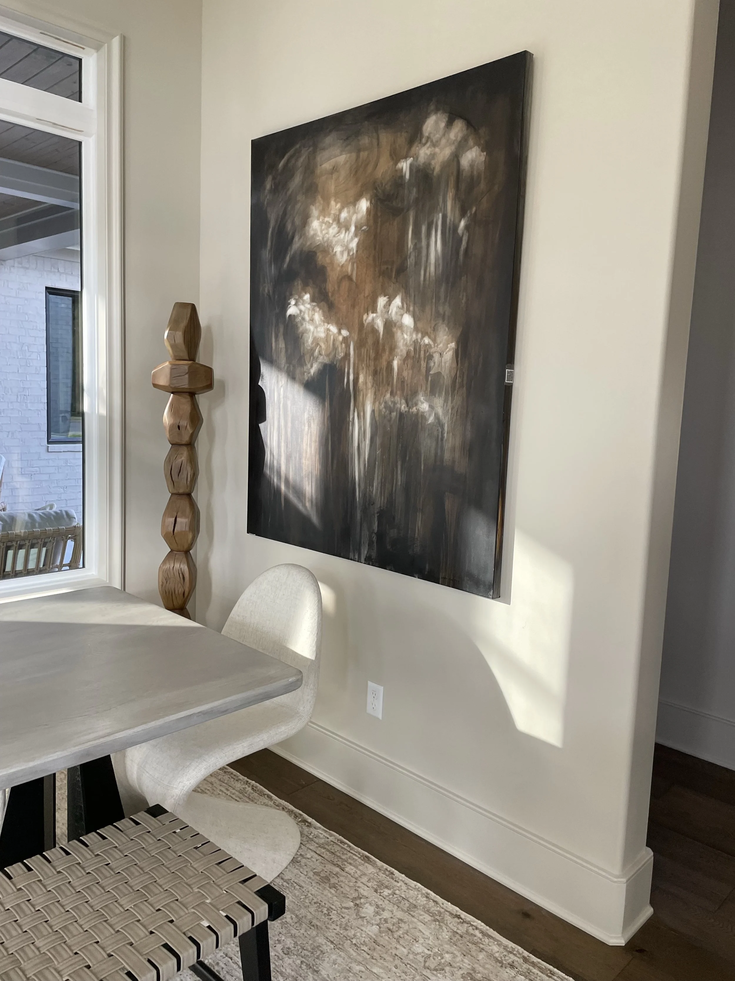 Interior of a room with a dining table, a light-colored upholstered chair, a woven bench, a large abstract painting on the wall, and a window showing an outdoor deck.