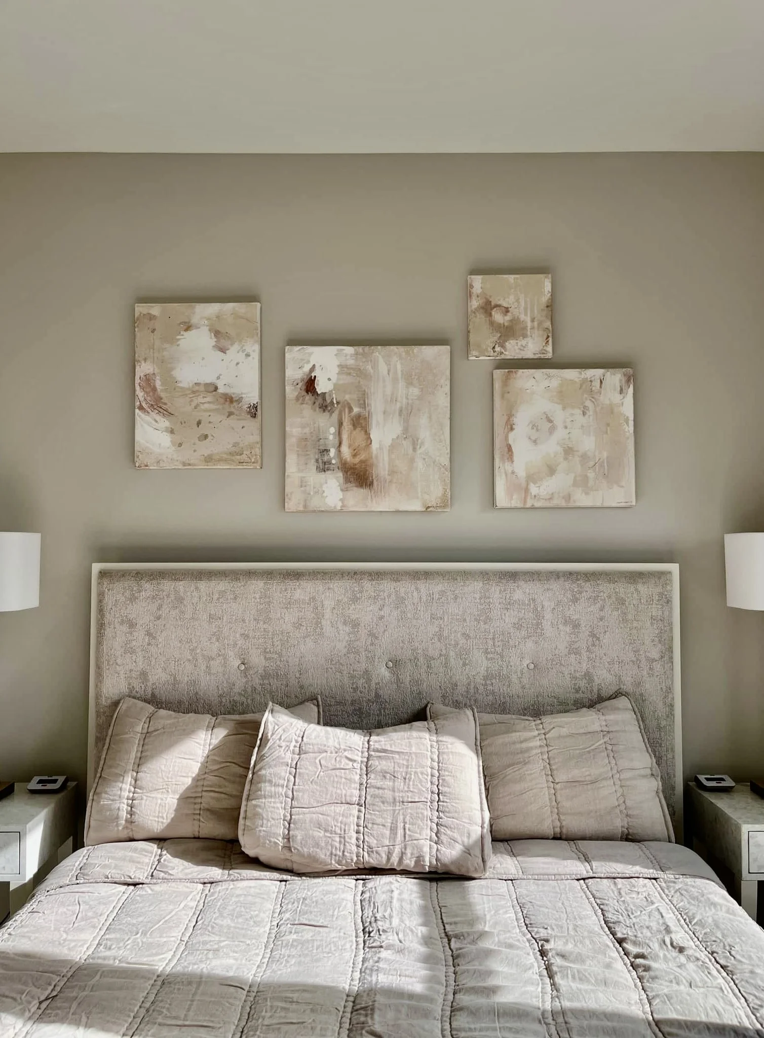 A neatly made bed with a beige quilt and matching pillow shams, flanked by two small white nightstands with lamps, against a light green wall decorated with four abstract artwork pieces.