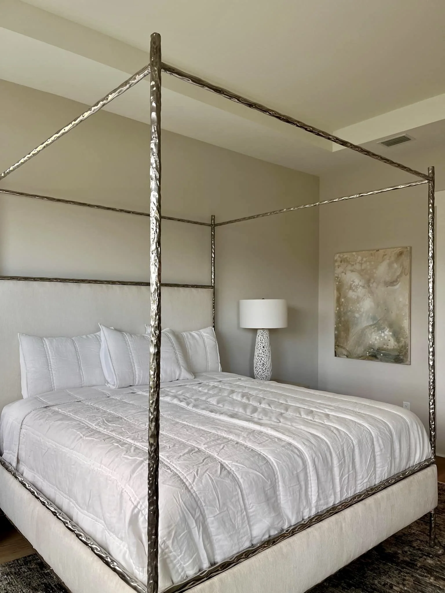 A bedroom with a four-poster bed frame made of textured silver metal, a white quilted bedspread, three white pillows, a white bedside lamp with a patterned shade, an abstract painting on the wall, and a dark patterned carpet.