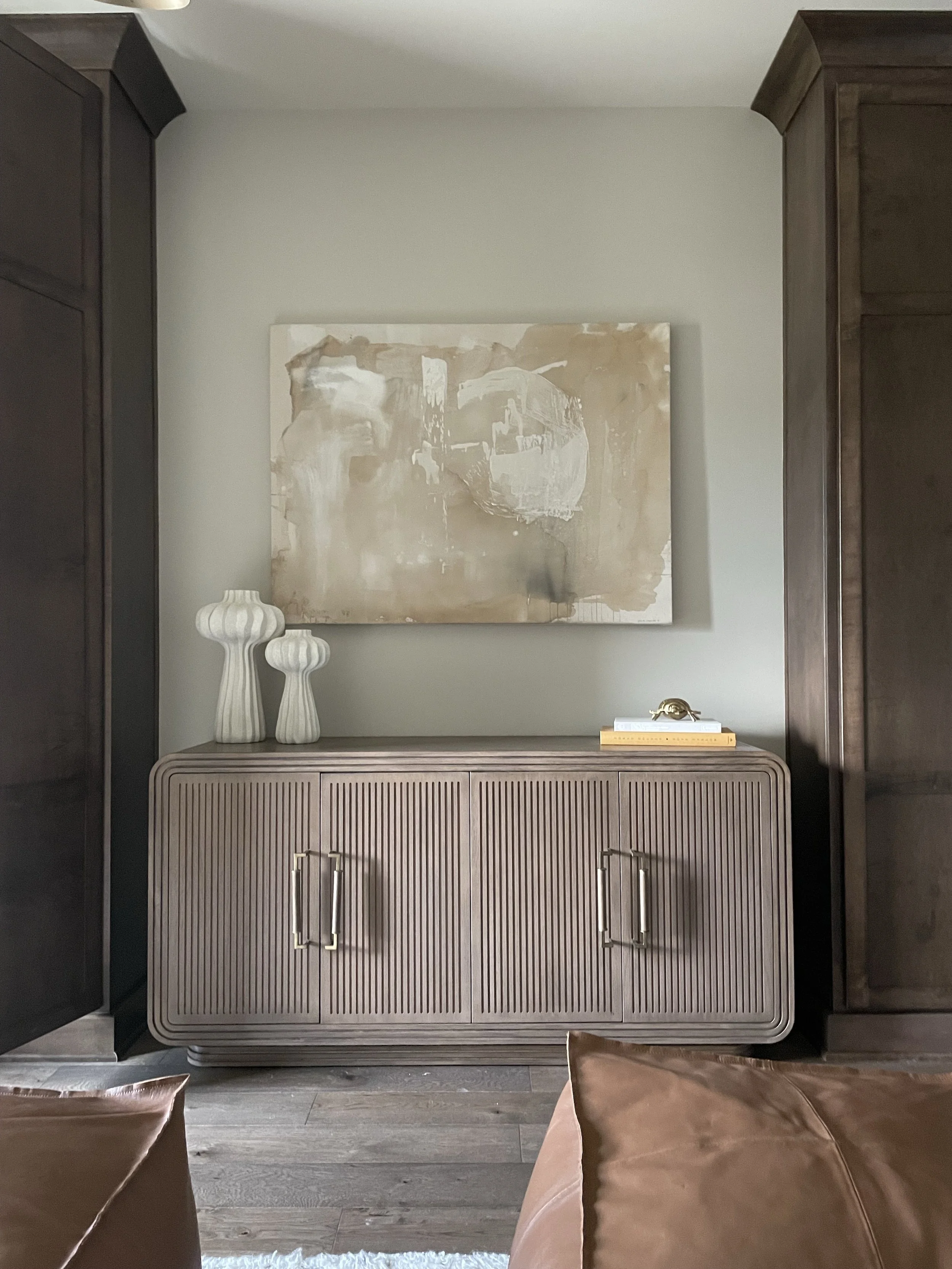 A beige wooden cabinet with vertical slats, gold handles, and rounded edges stands against a light-colored wall. Above it hangs a large abstract painting in neutral tones. To the left are two tall, white ceramic vases with ribbed textures. To the rig
