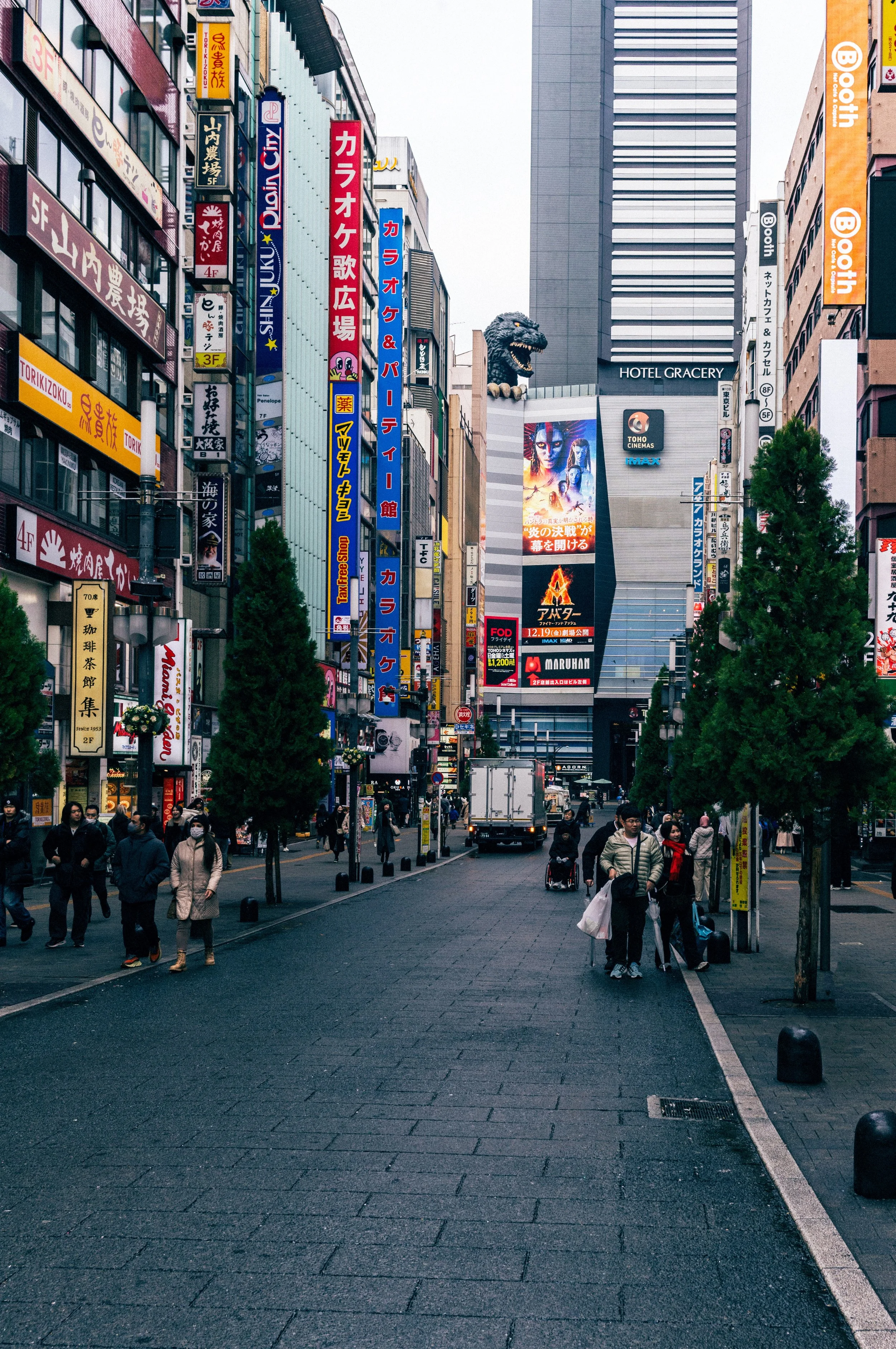 Busy Shinjuku street scene with neon signs, pedestrians, and the Godzilla head visible above Hotel Gracery Tokyo.