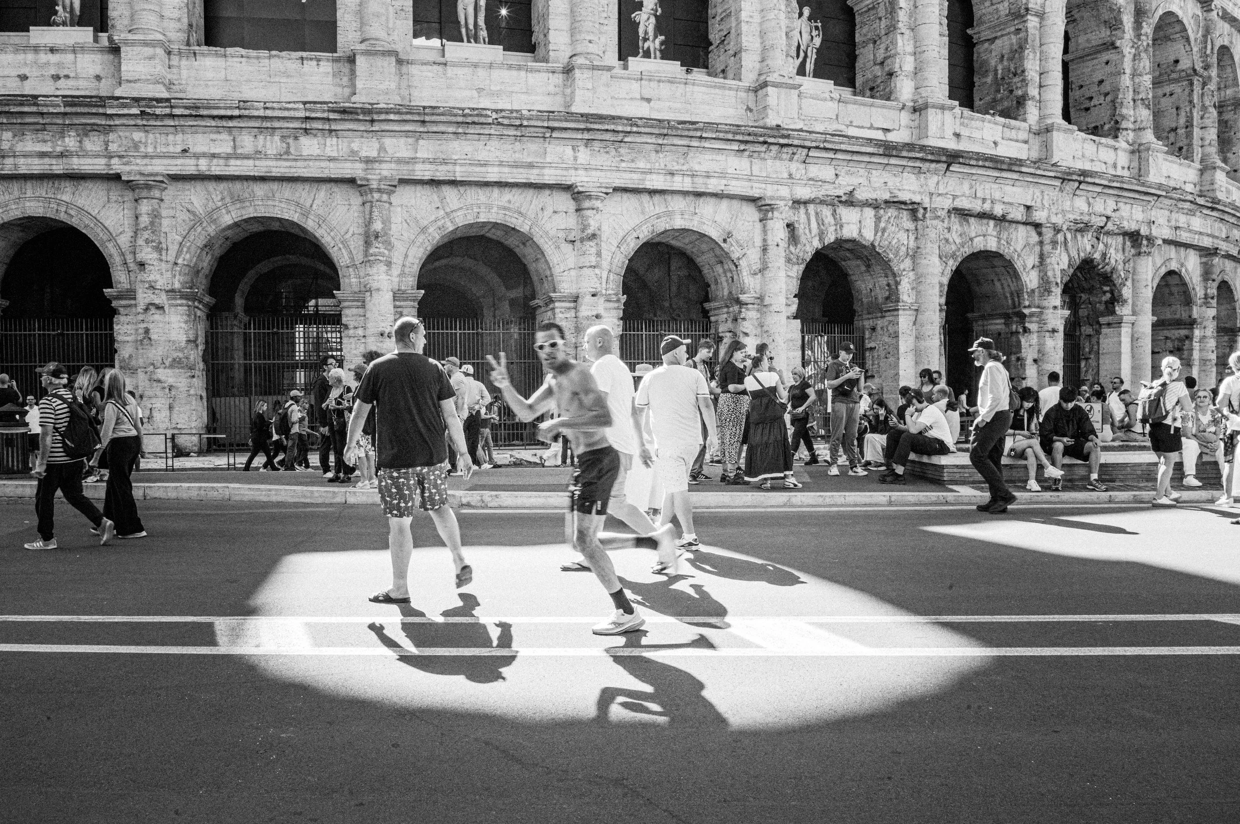 A runner flashes a peace sign while crossing the road outside the Colosseum in Rome, captured in a candid black and white street photograph.