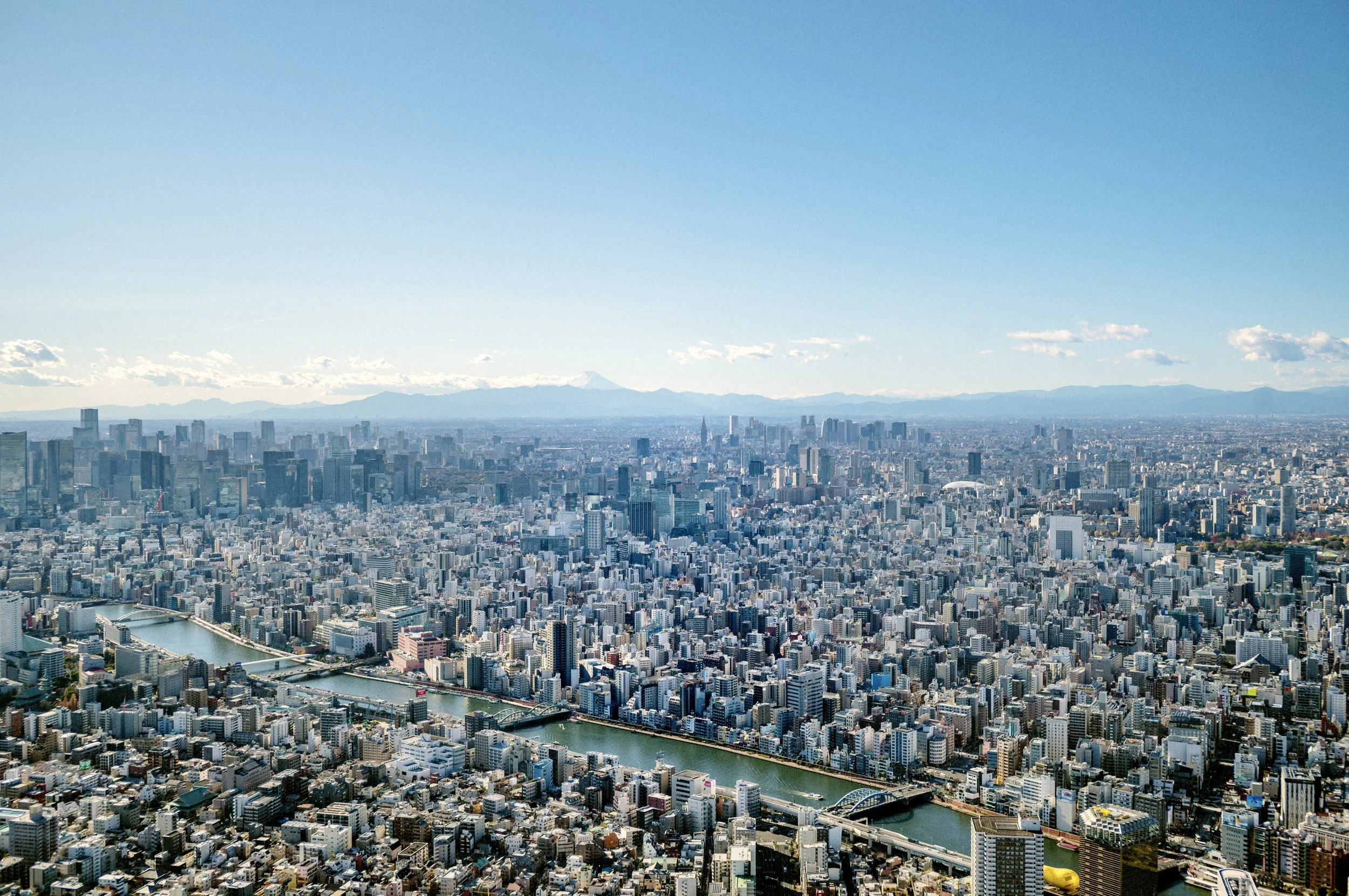 Wide aerial view of Tokyo skyline with rivers, dense city buildings, and Mount Fuji visible on the horizon.