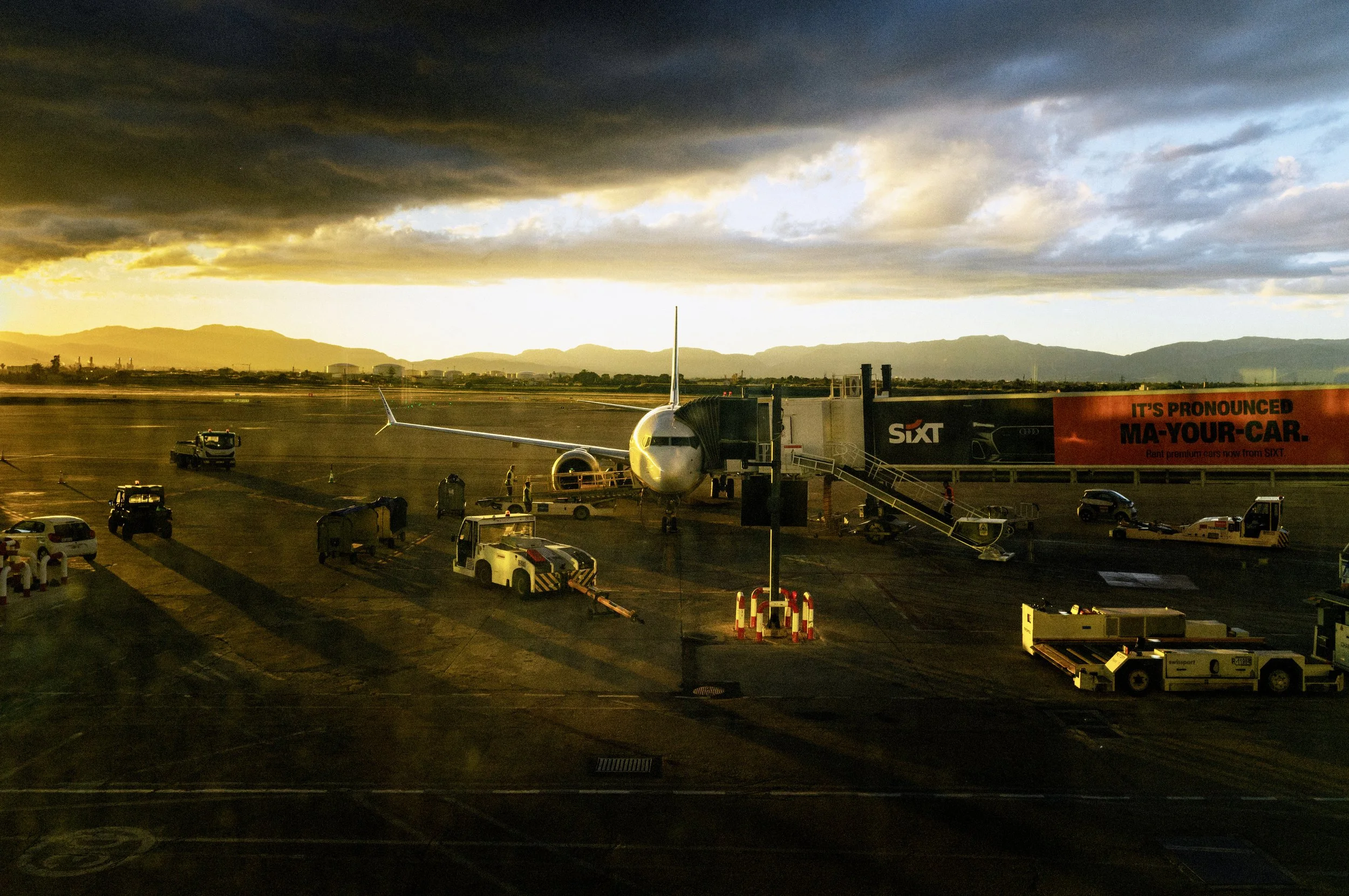 Commercial aircraft parked at the gate at Palma de Mallorca Airport beneath dramatic storm clouds as golden sunset light illuminates the runway and the Tramuntana mountains in the distance. Optional Image Title