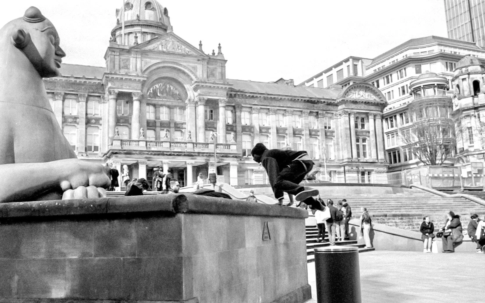A man leaps onto the stone plinth beside the Birmingham Town Hall sphinx in a spontaneous black and white street photograph.