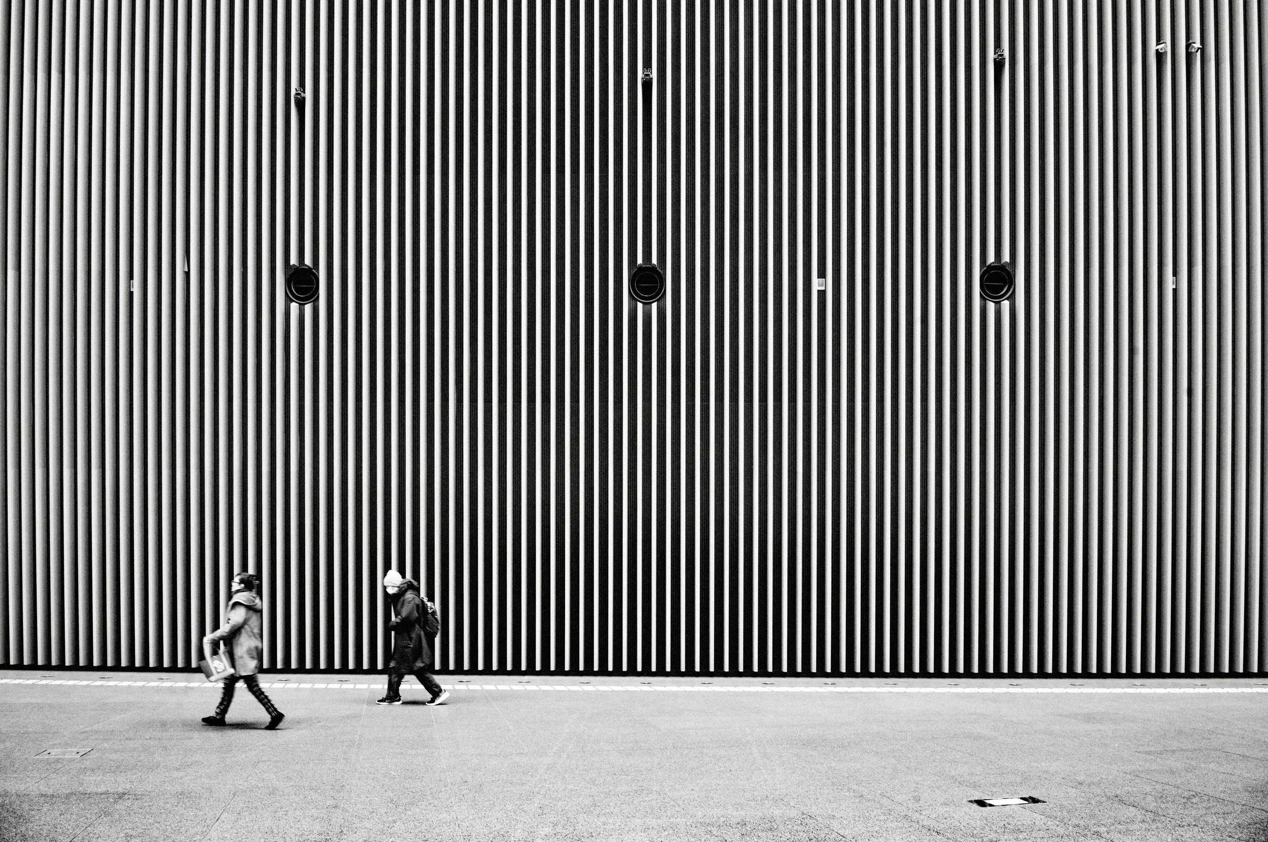 Two people walking past a striped modern wall in Tokyo captured in black and white minimalist street photography style.