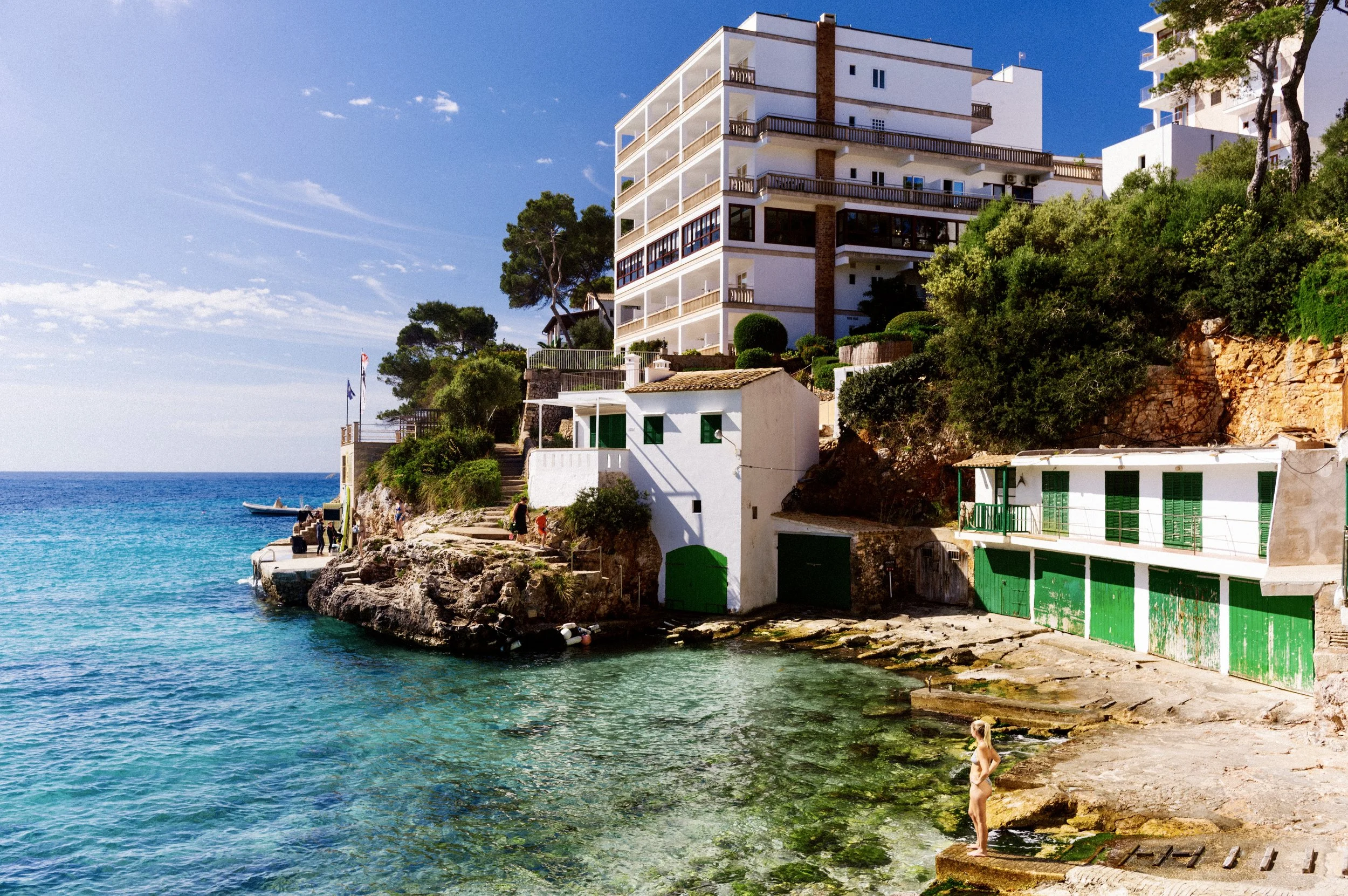 Whitewashed house with green shutters above a rocky swimming cove with clear blue water in Mallorca