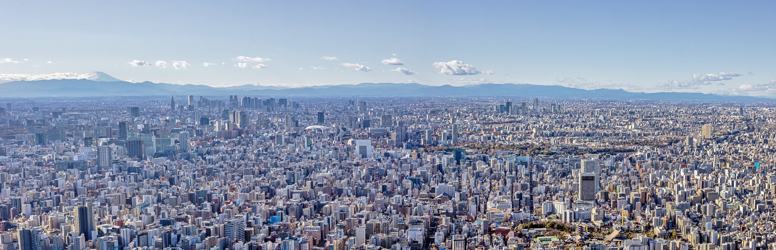Ultra-wide panoramic photograph of Tokyo city skyline under a bright blue sky with distant mountains beyond the urban landscape.