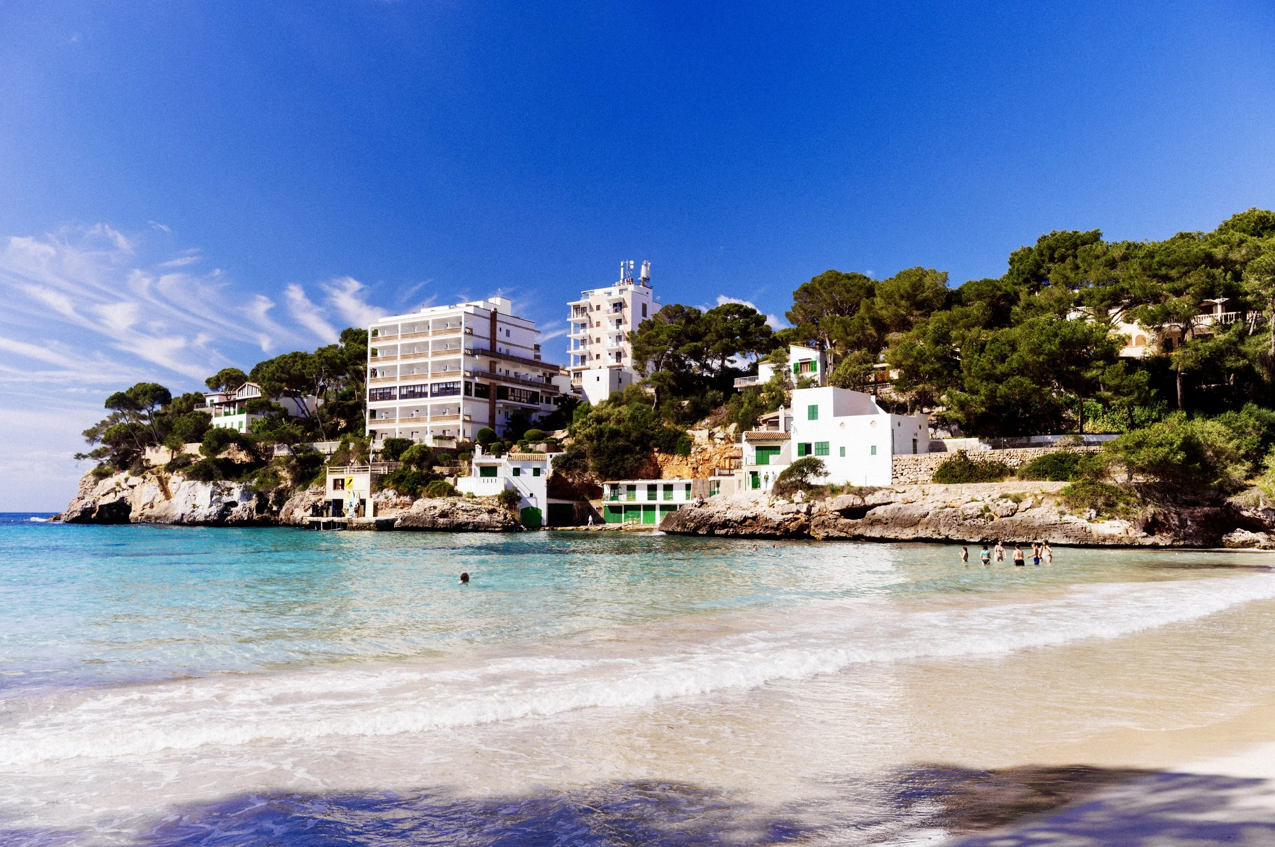 Gentle waves on the white sand of Cala Santanyí beach with cliffside buildings, Mallorca
