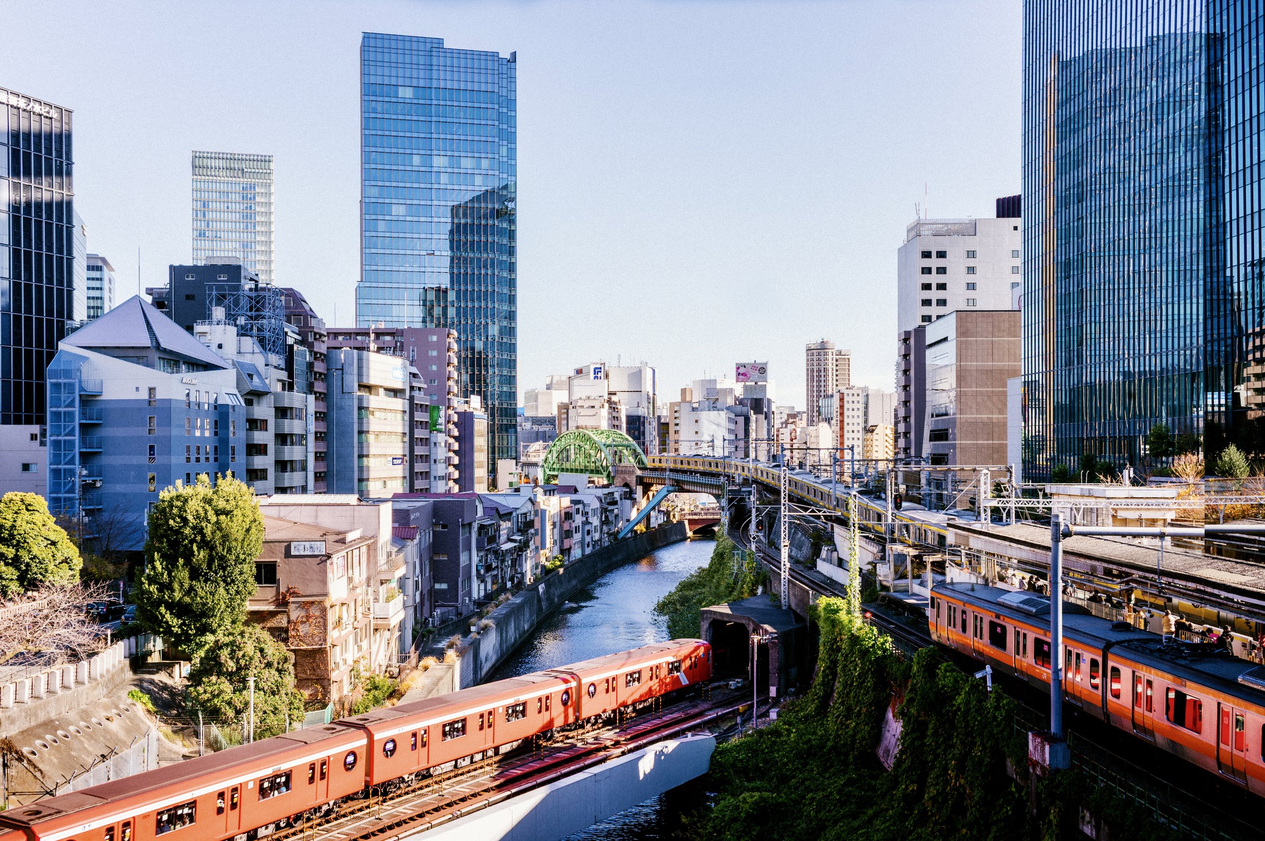 Multiple Tokyo trains crossing over rivers and tracks near Hijiri-bashi Bridge surrounded by city buildings.