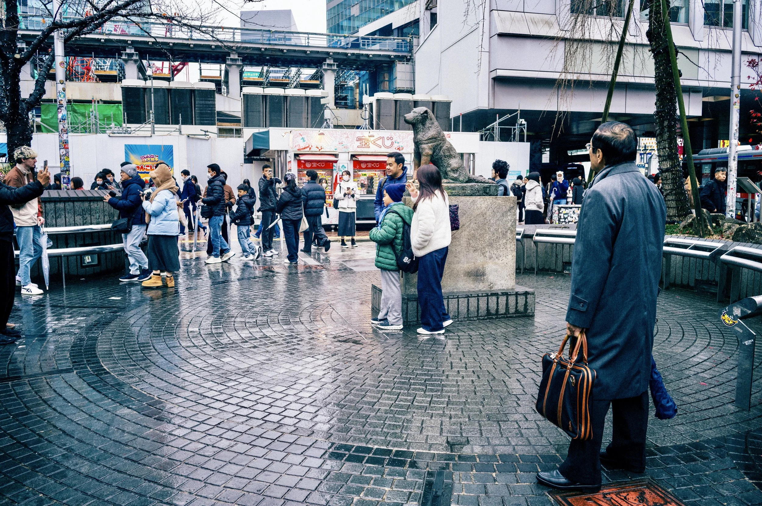People gathering around the Hachikō Statue near Shibuya Station on a rainy day in Tokyo.