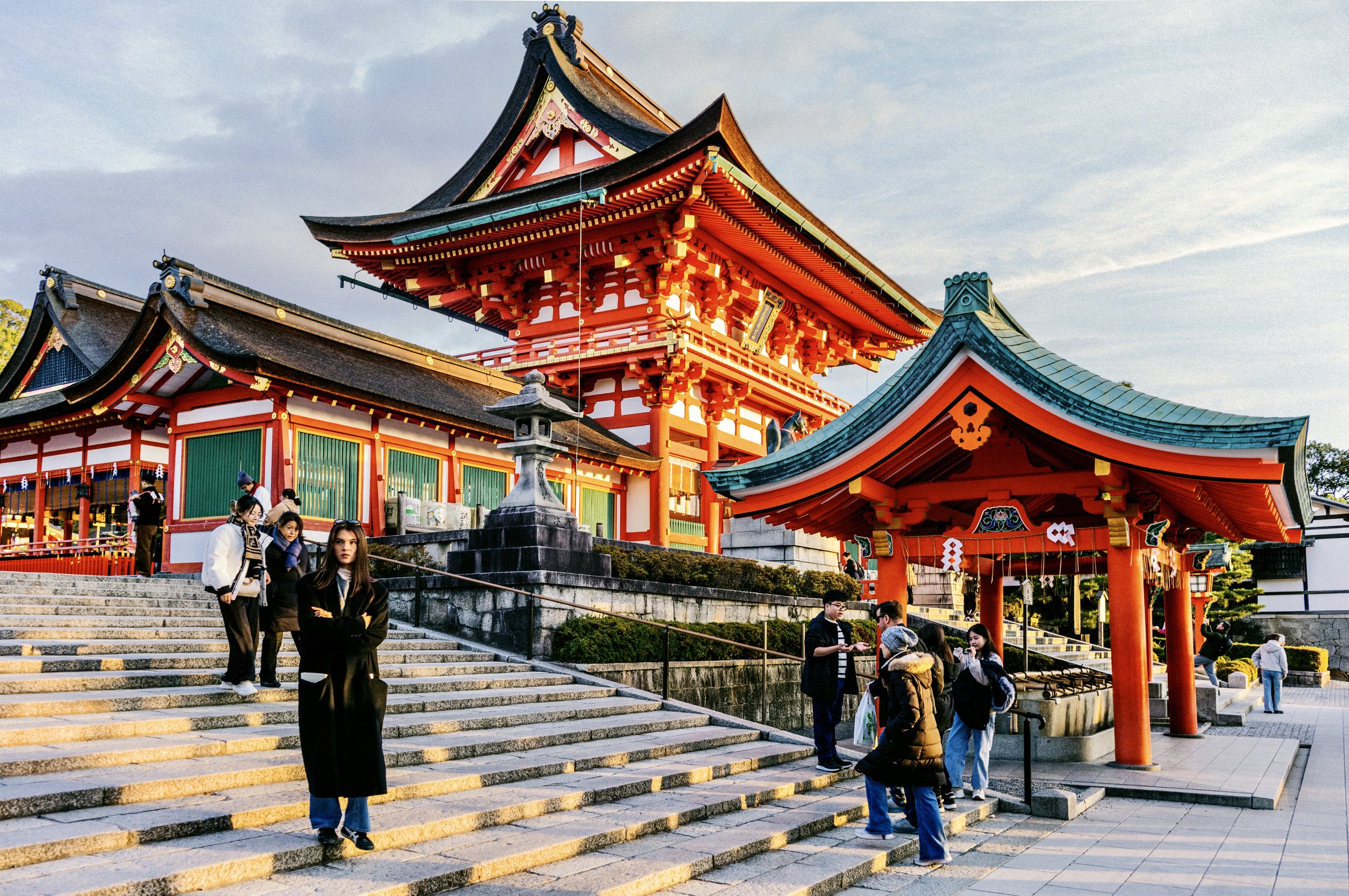 Crowds gathering at the ornate main shrine building of Fushimi Inari Taisha in Kyoto during golden hour.